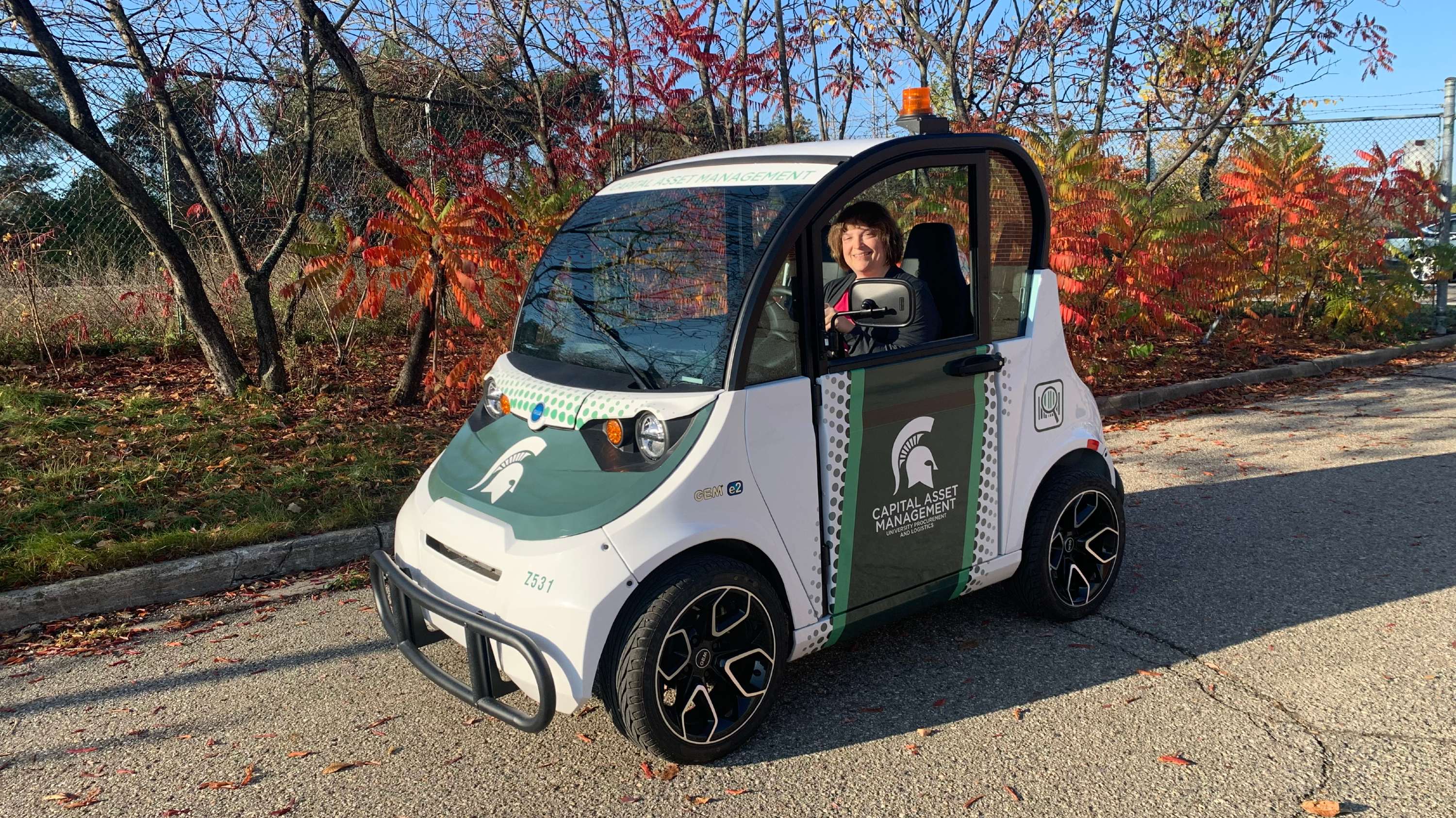 CAM Office Assistant Susan Bauer drives a green and white electric car with MSU decals down road with red and orange fall leaves in the background.