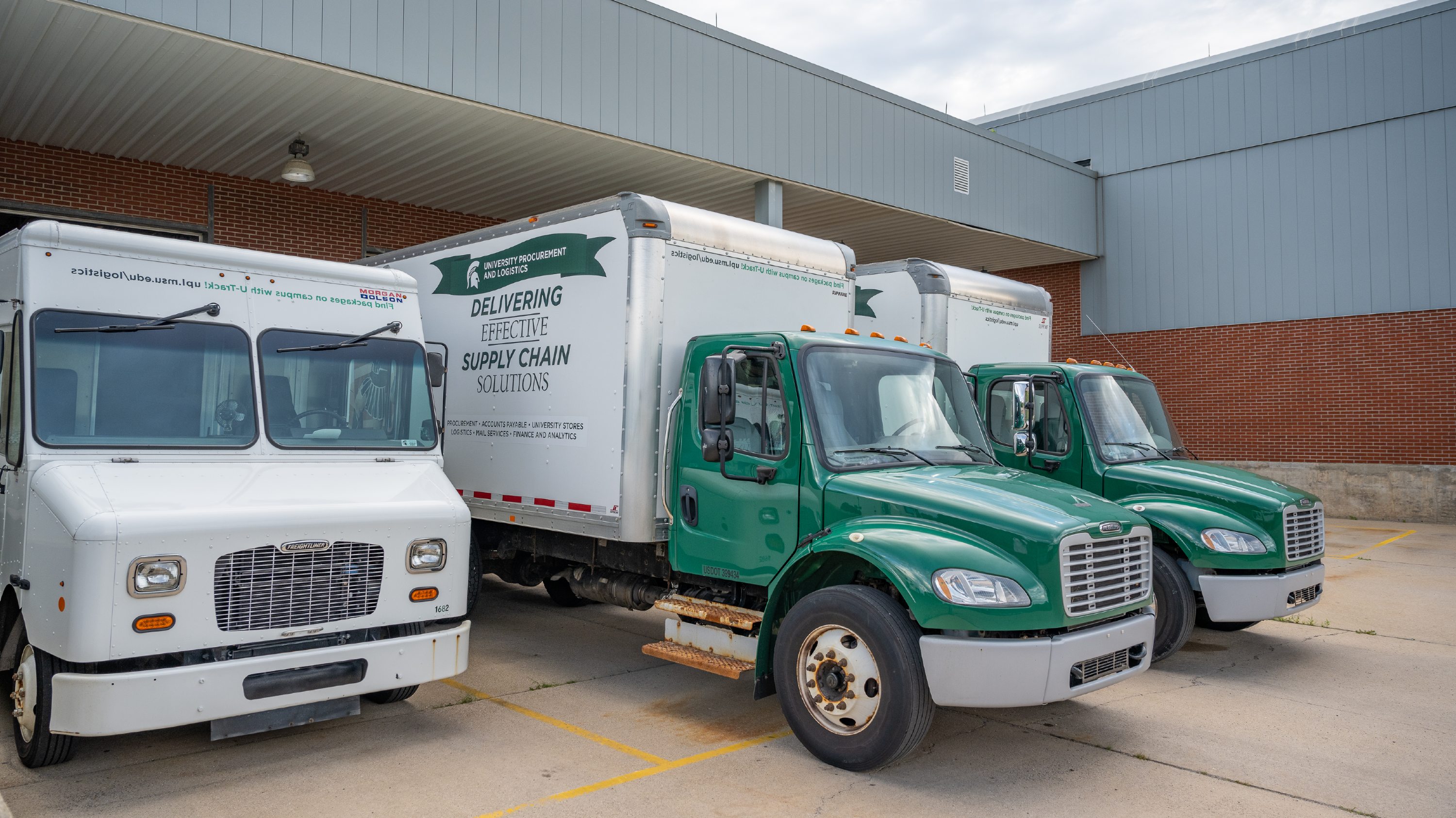 Three trucks facing right are backed into a loading dock. The first truck on the left is all white. The other two trucks have a green cab and white trailers.