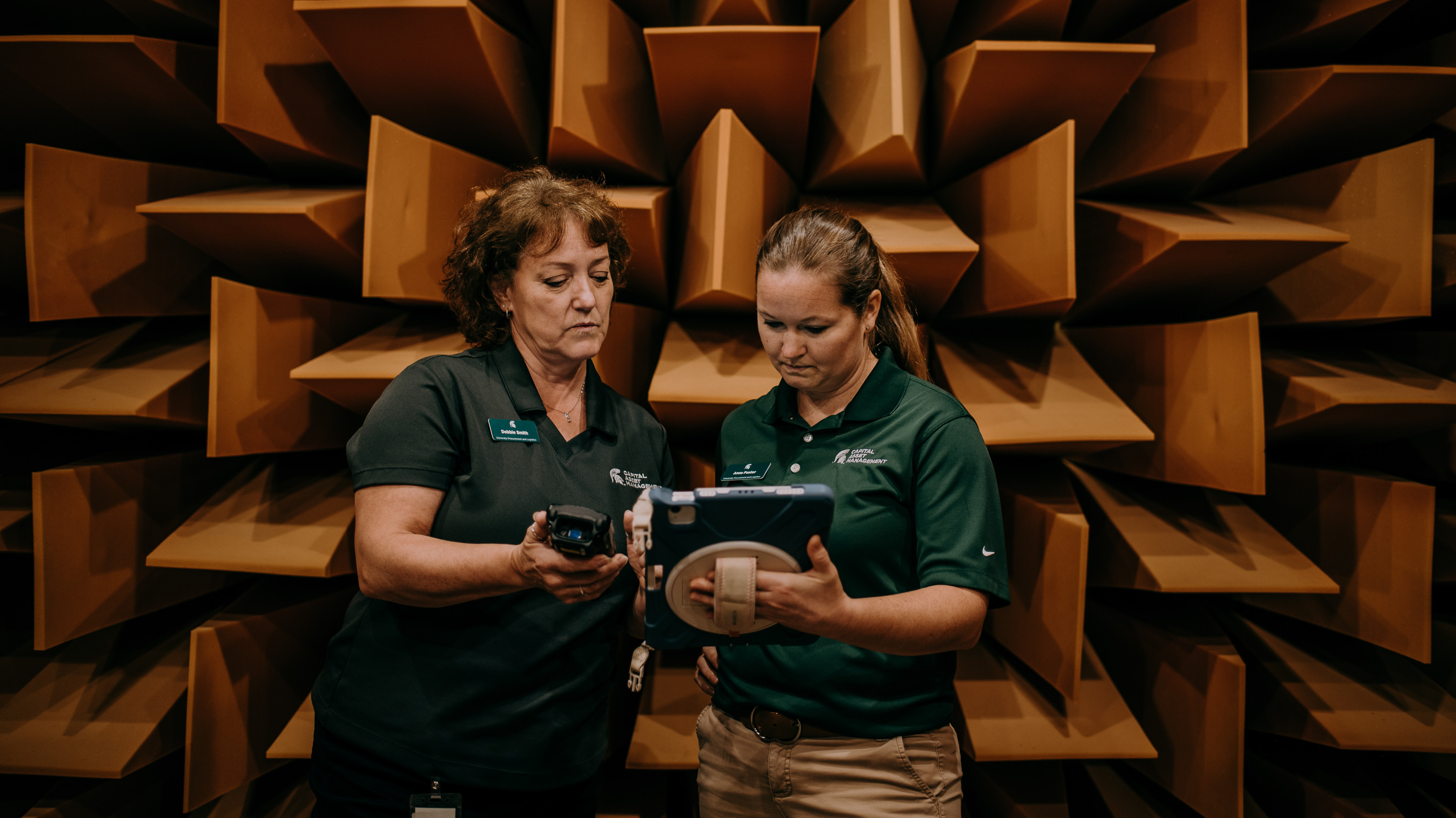 Debbie Smith and Anna Foster stand together looking at a CAM tablet inside an anechoic chamber with rusty orange foam sound absorbers jutting out from the wall behind them.