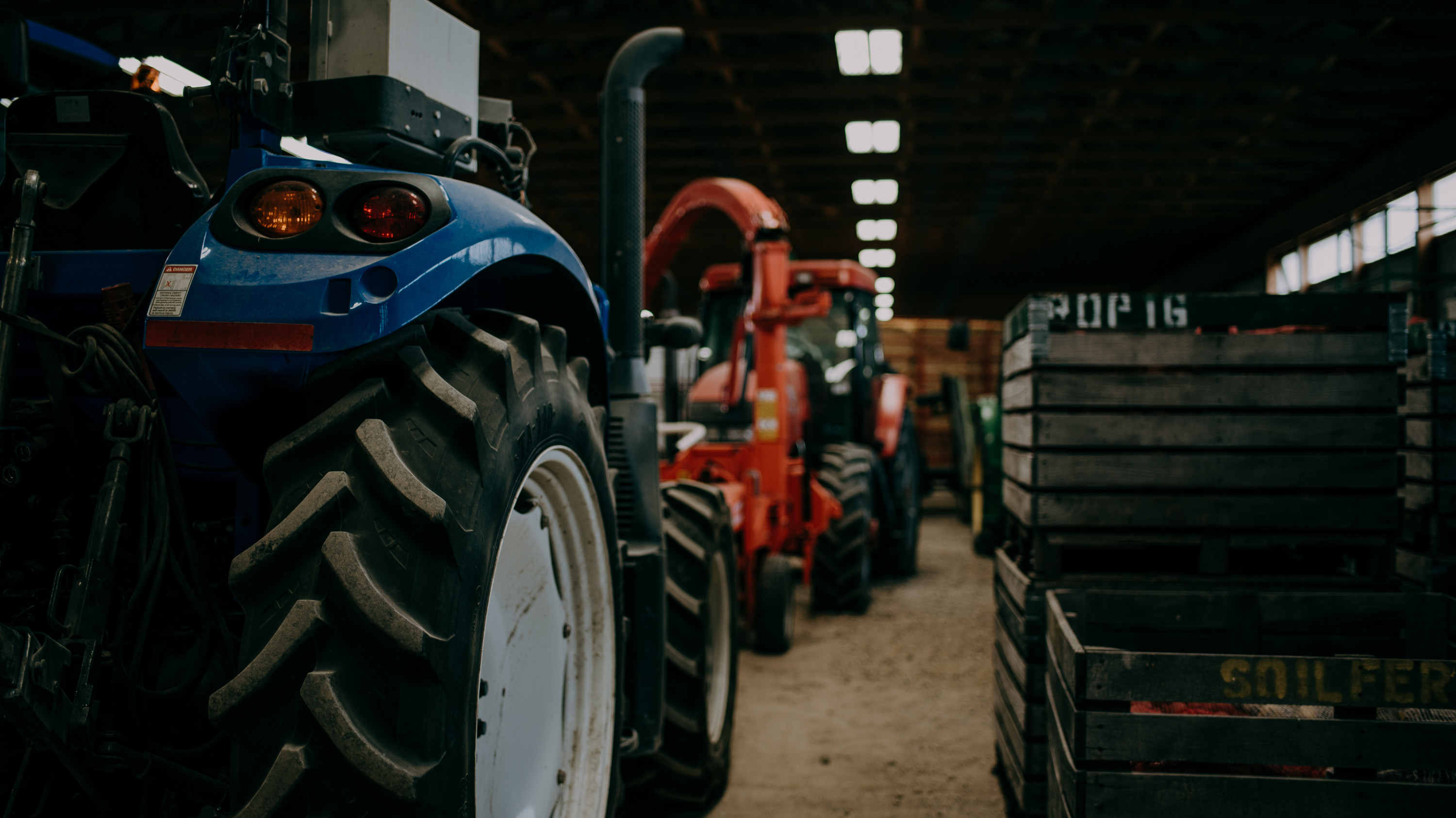 One blue and one red tractor, both with large black tires, are parked near multiple stacked wooden crates inside a barn.