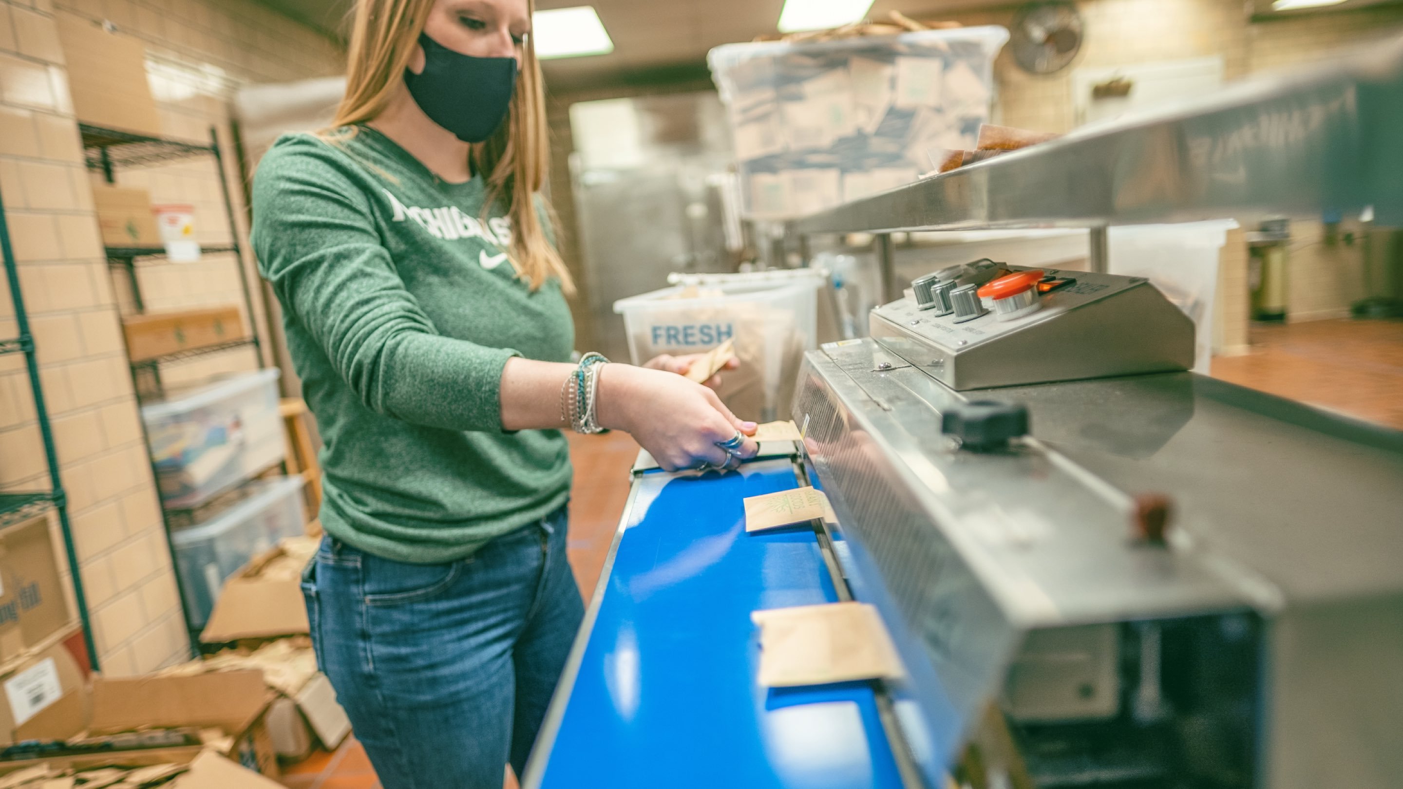 A female with bold hair in a MSU t-shirt and wearing a face covering inserts tea packets into a machine for closure.