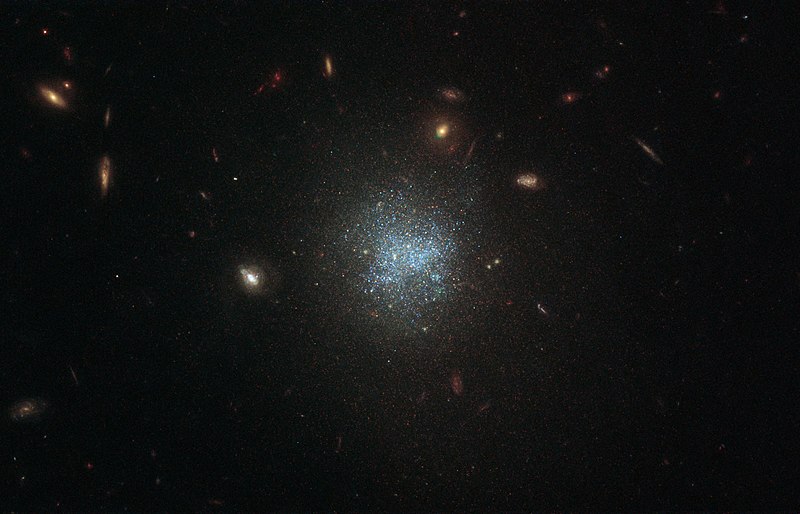 A dark telescope image is dotted with small, yellow spiral and disc-shaped galaxies. At its center, though, the galaxy shown looks like a dispersed cloud of blue dots.
