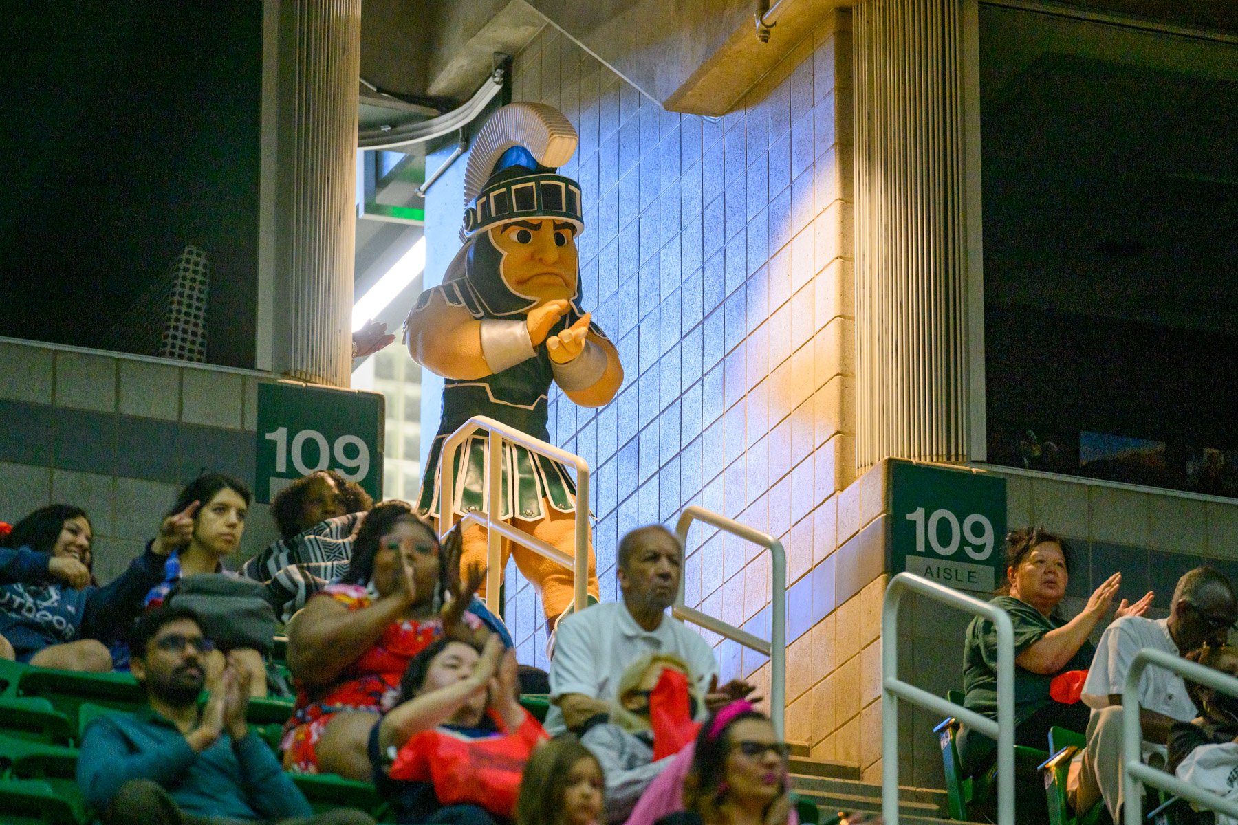 Sparty applauds at the top of the Breslin Center's arena leading to the concourse area with members of the audience sitting in the foreground.