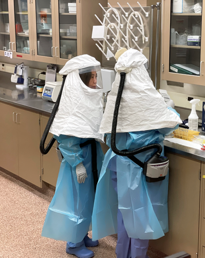 Wise and Thompson handle samples to prepare for testing. Note that both are wearing powered air-purifying respirators and other personal protective equipment, including shoes, that are only used in this space and will be thoroughly disinfected following completion of testing.