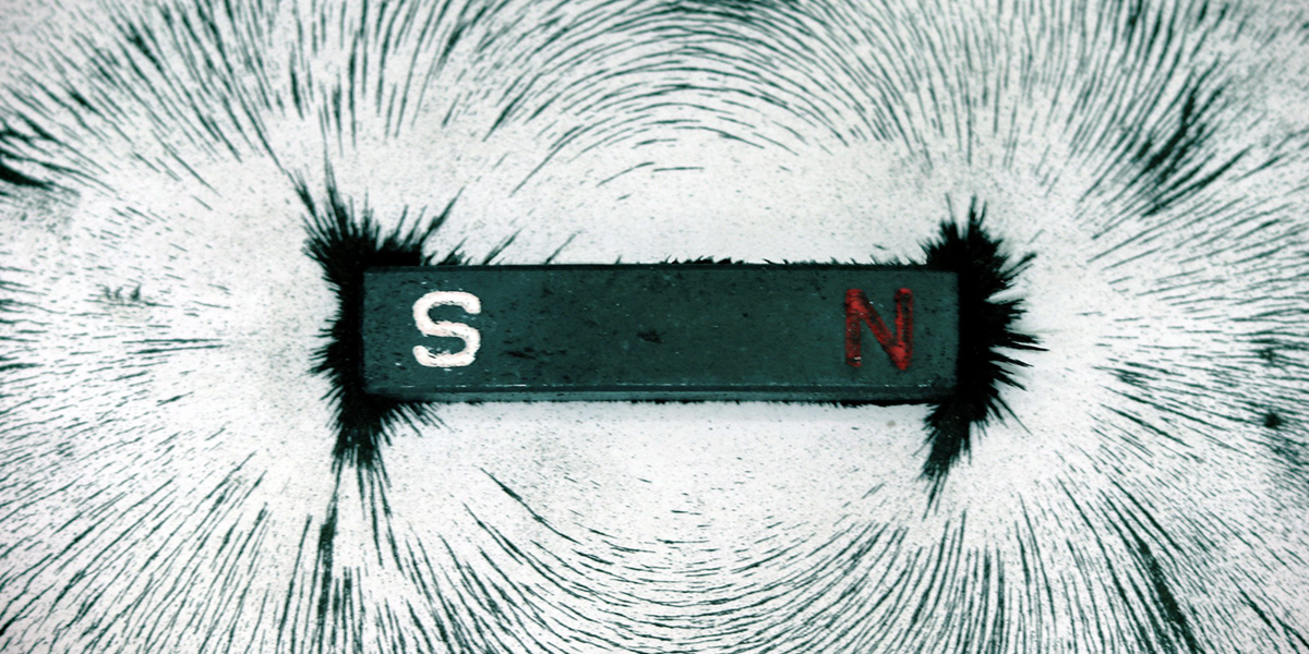 A photograph shows a gray bar magnet labeled with a white “S” and red “N” (for the south and north poles, respectively) is surrounded by metal filings arranged in dark loops around the magnet.