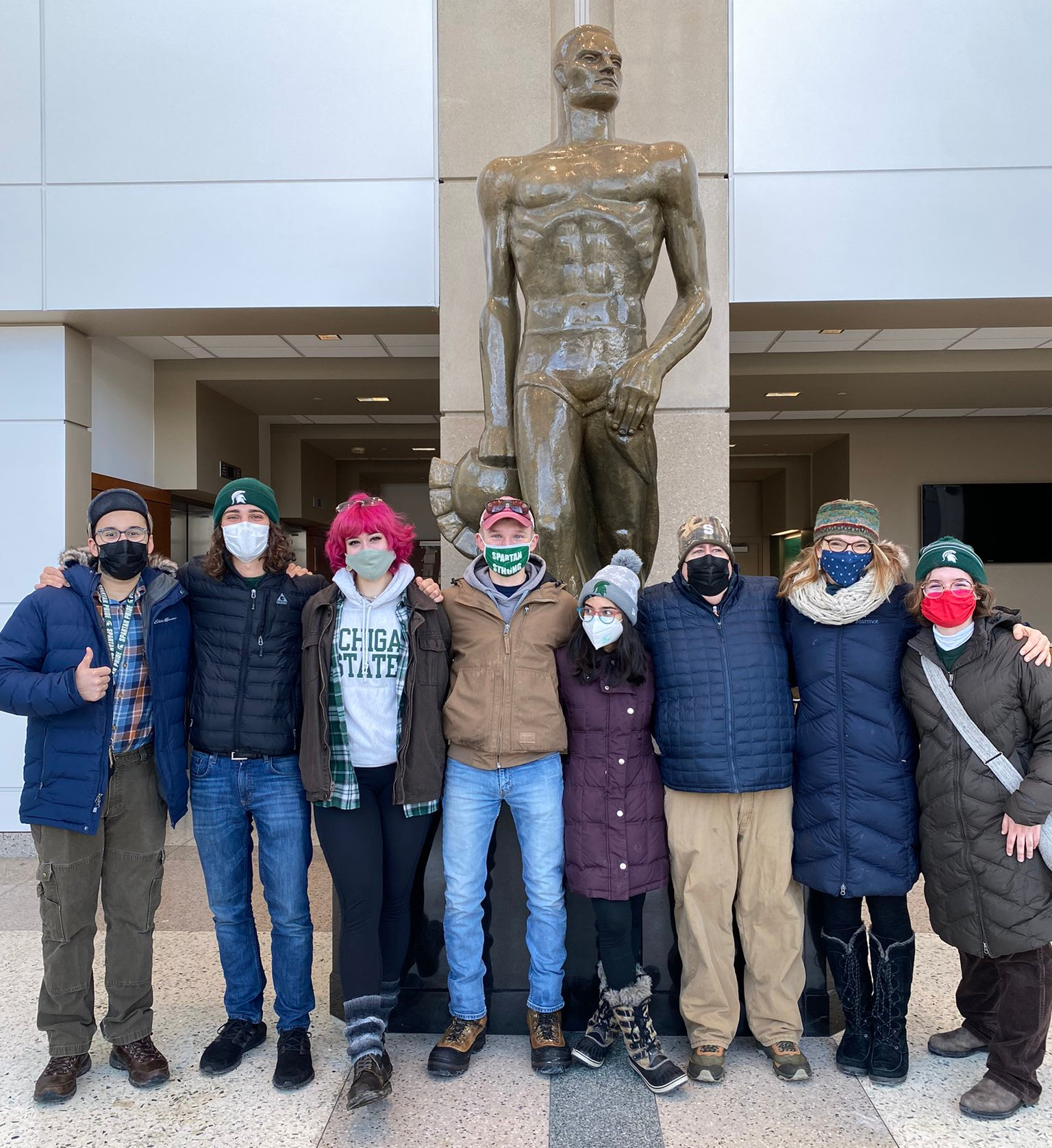 Eight people in winter jackets and masks stand in front of the Spartan statue in Spartan Stadium.