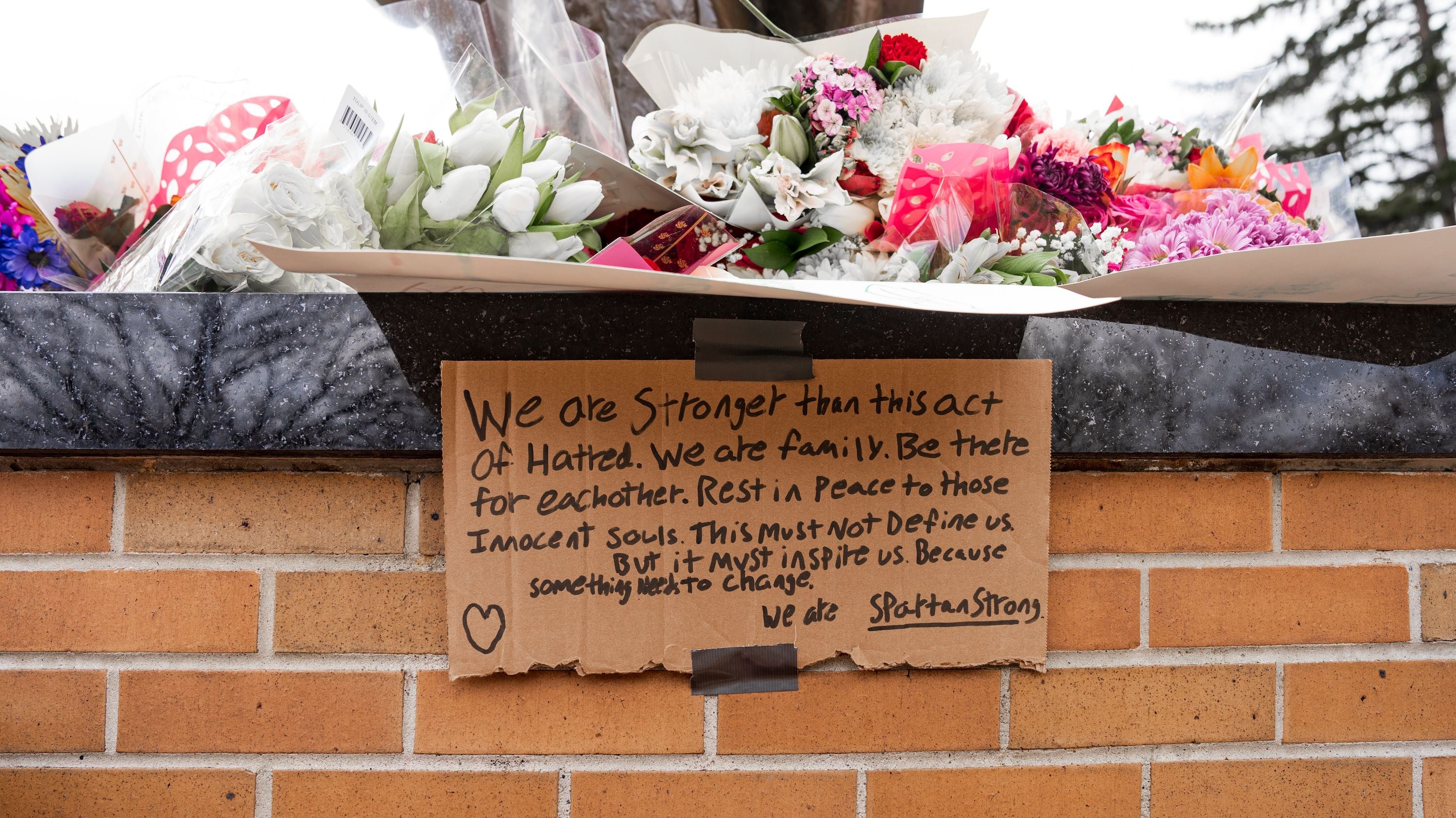 A handwritten sign beneath flowers at the Spartan Statue that reads: We are stronger than this act of hatred. We are family. Be there for each other. Rest in peace to those innocent souls. This must not define us. But it must inspire us. Because something needs to change. We are Spartan Strong.