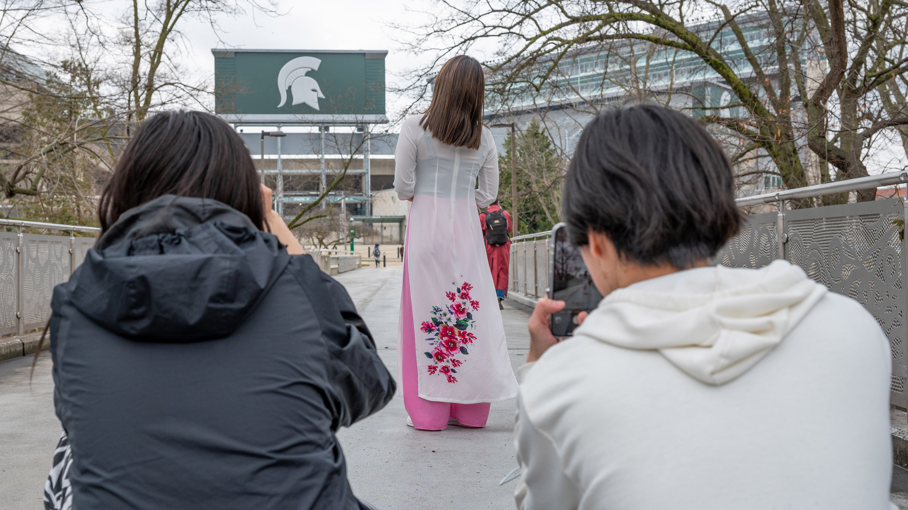 Tw students taking pictures. of student wearing traditional Vietnamese dress, on MSU library bridge