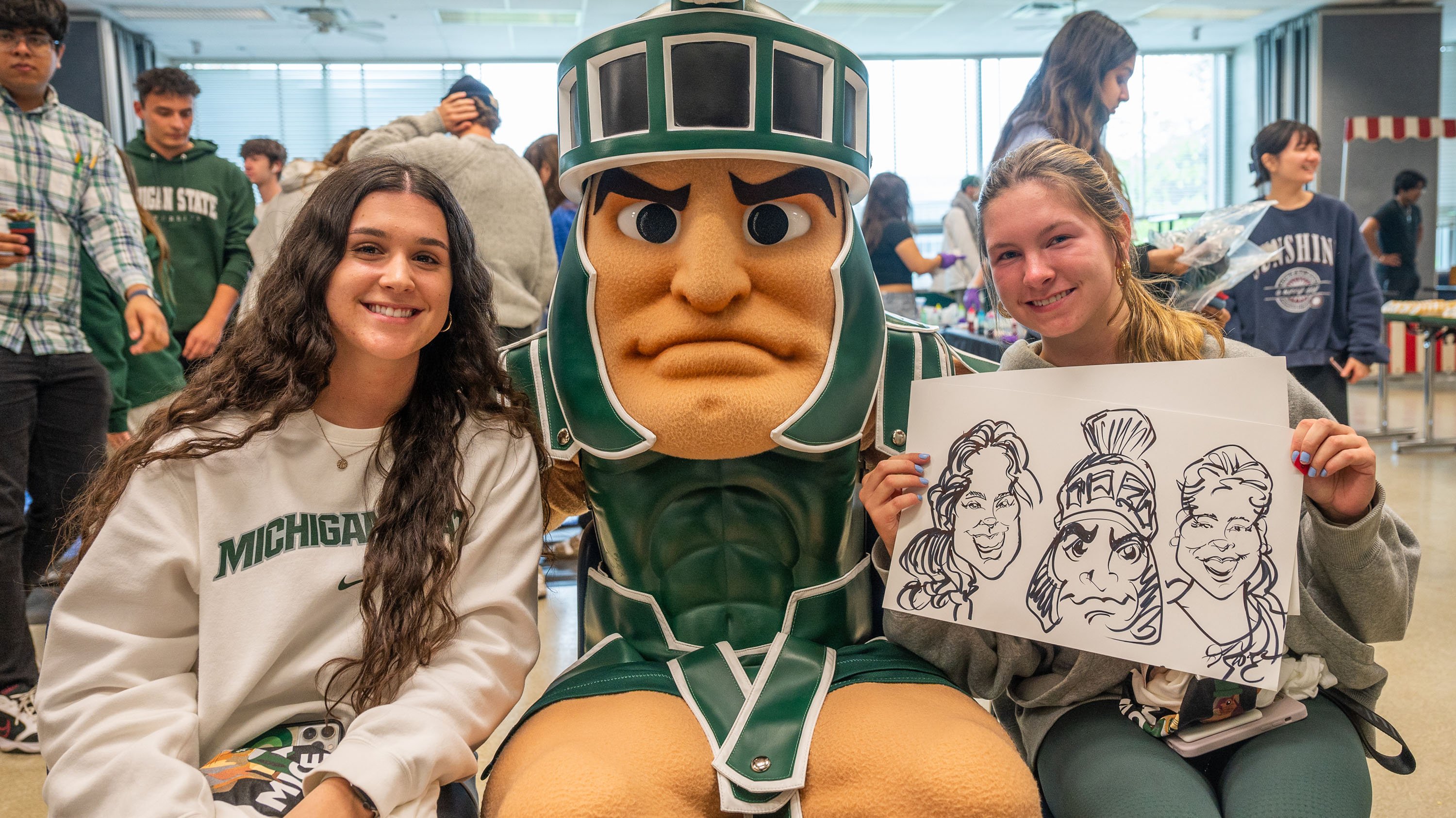 Two students posing with Sparty and a caricature drawing