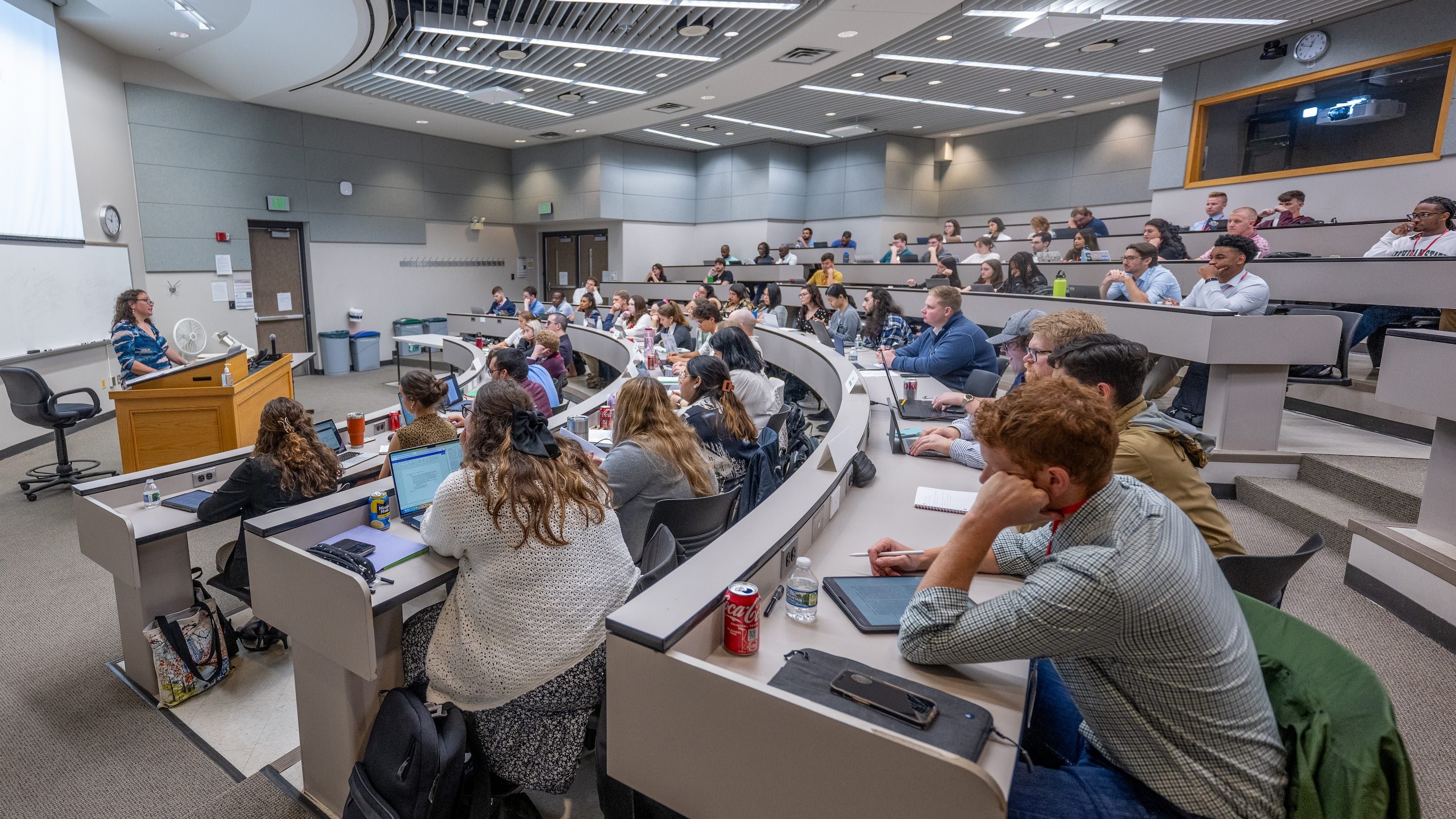 A lecturer speaks to a group of students in a large hall with stadium-style seating