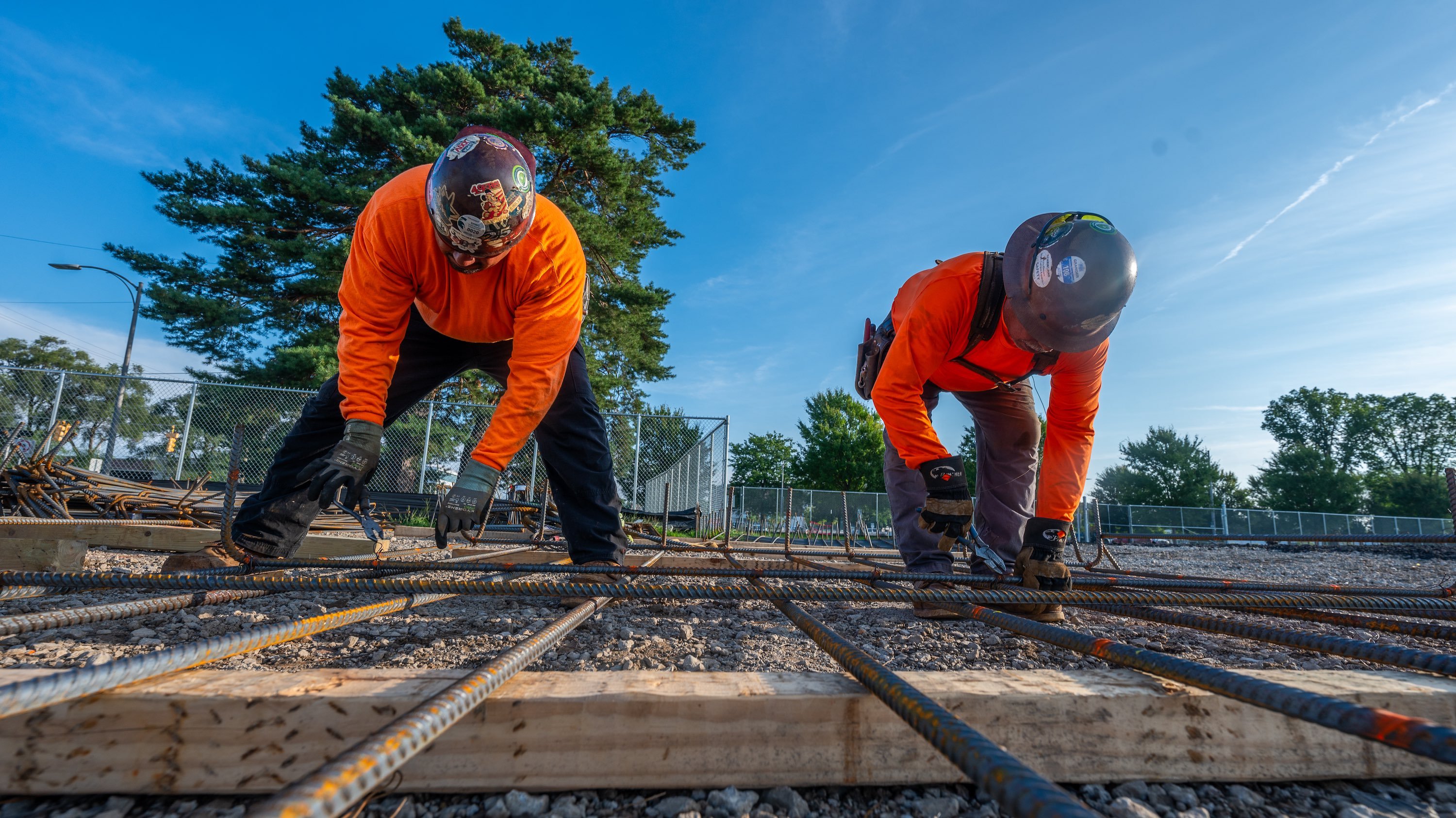Two construction workers in orange shirts and helmets work on a row of wood and steal rods