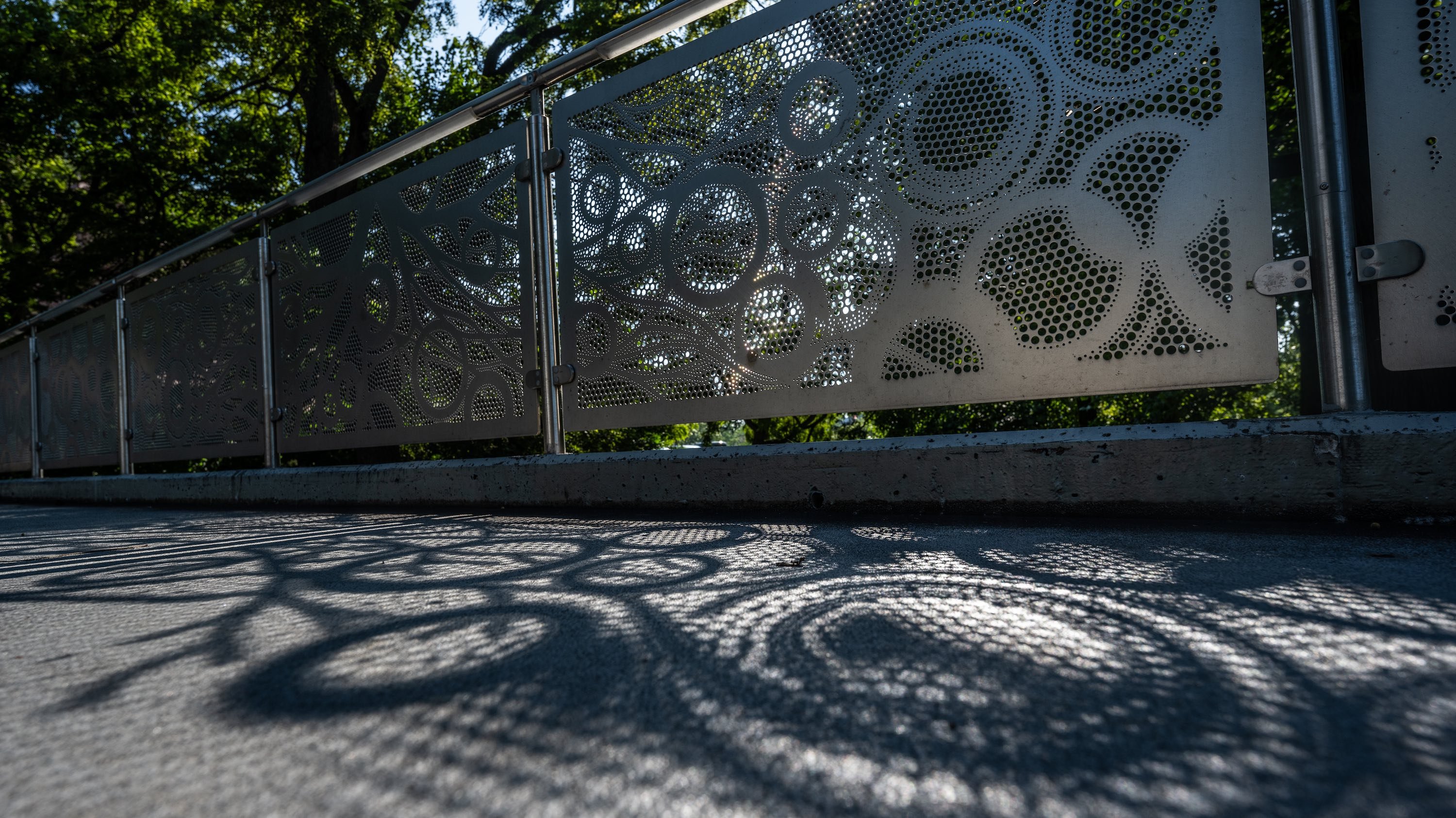 Sun peaks through panels that line a bridge, leaving a shadow of dots and circles swishing along the ground.