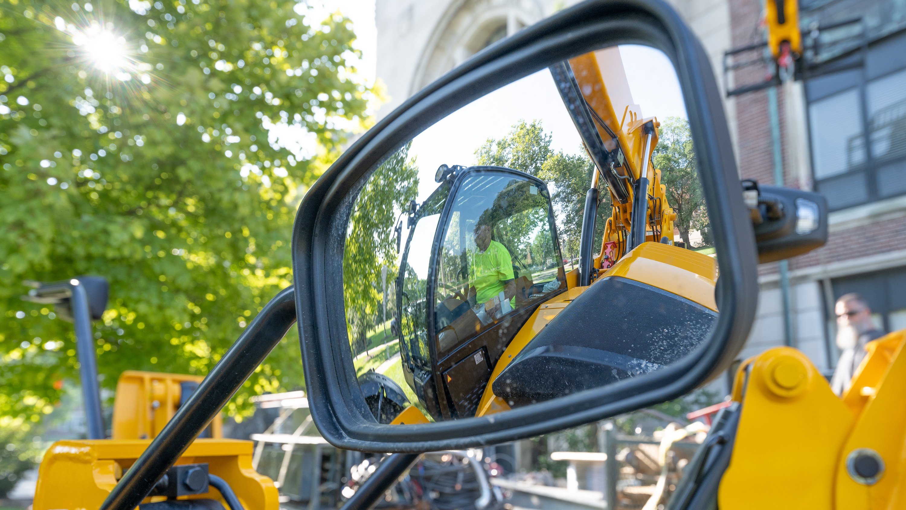 A view of a side-view mirror that looks into a yellow tractor, with a worker in a neon yellow shirt driving the machine.