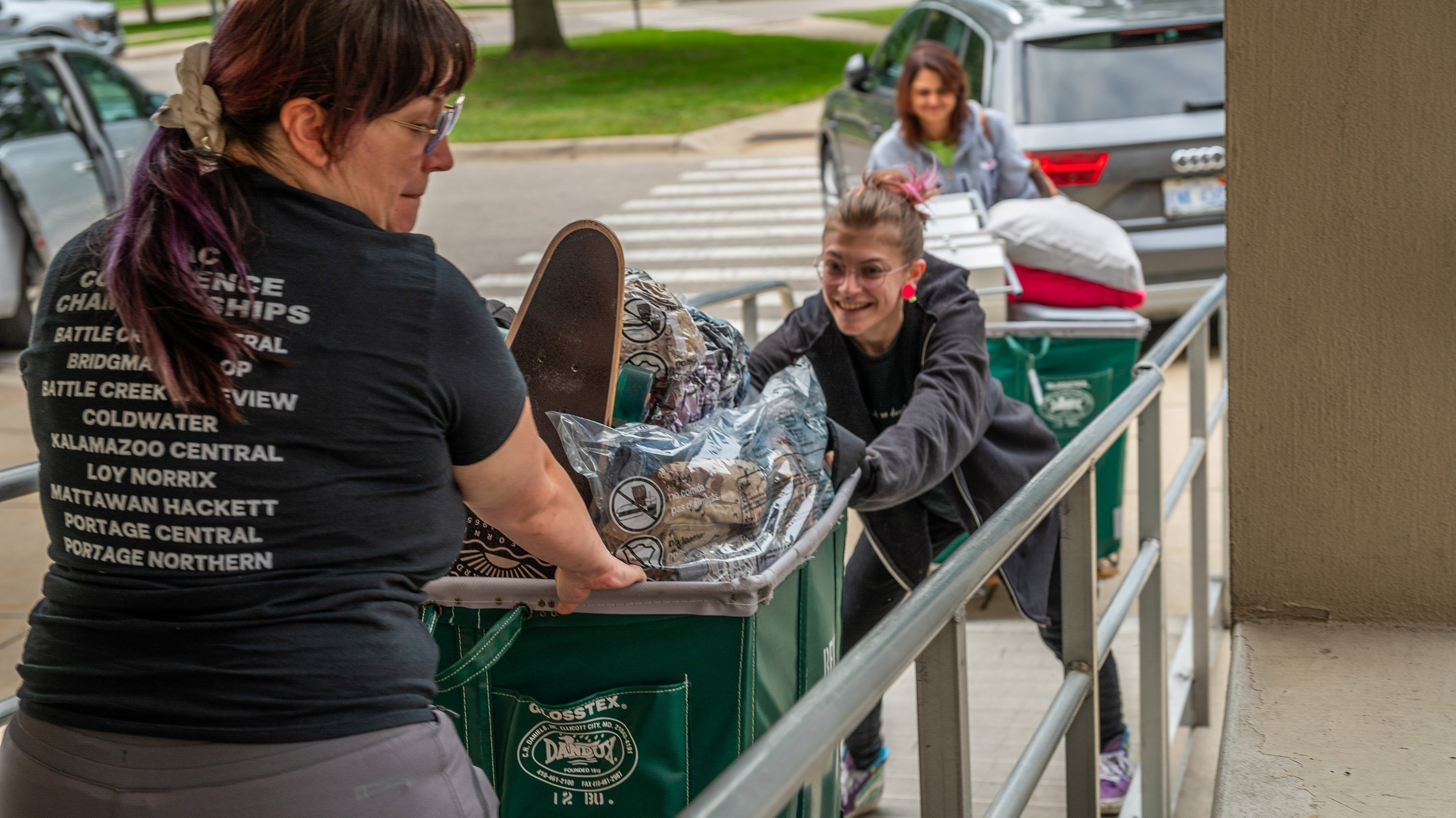 Three people pushing a cart filled with luggate up a ramp into a residence hall