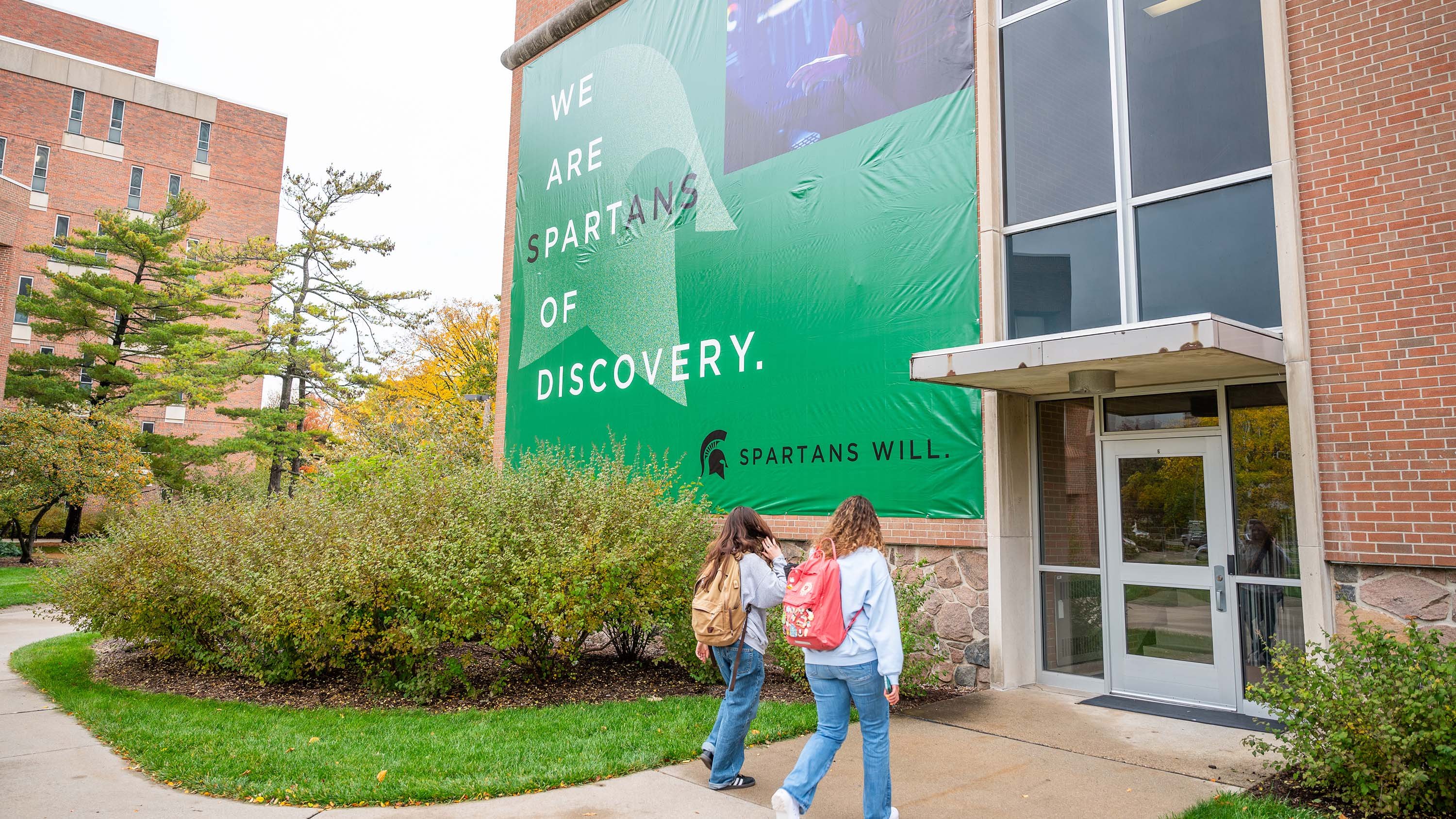 Large banner with text 'we are Spartans of Discovery' on entrnace of International Center, with students walking in
