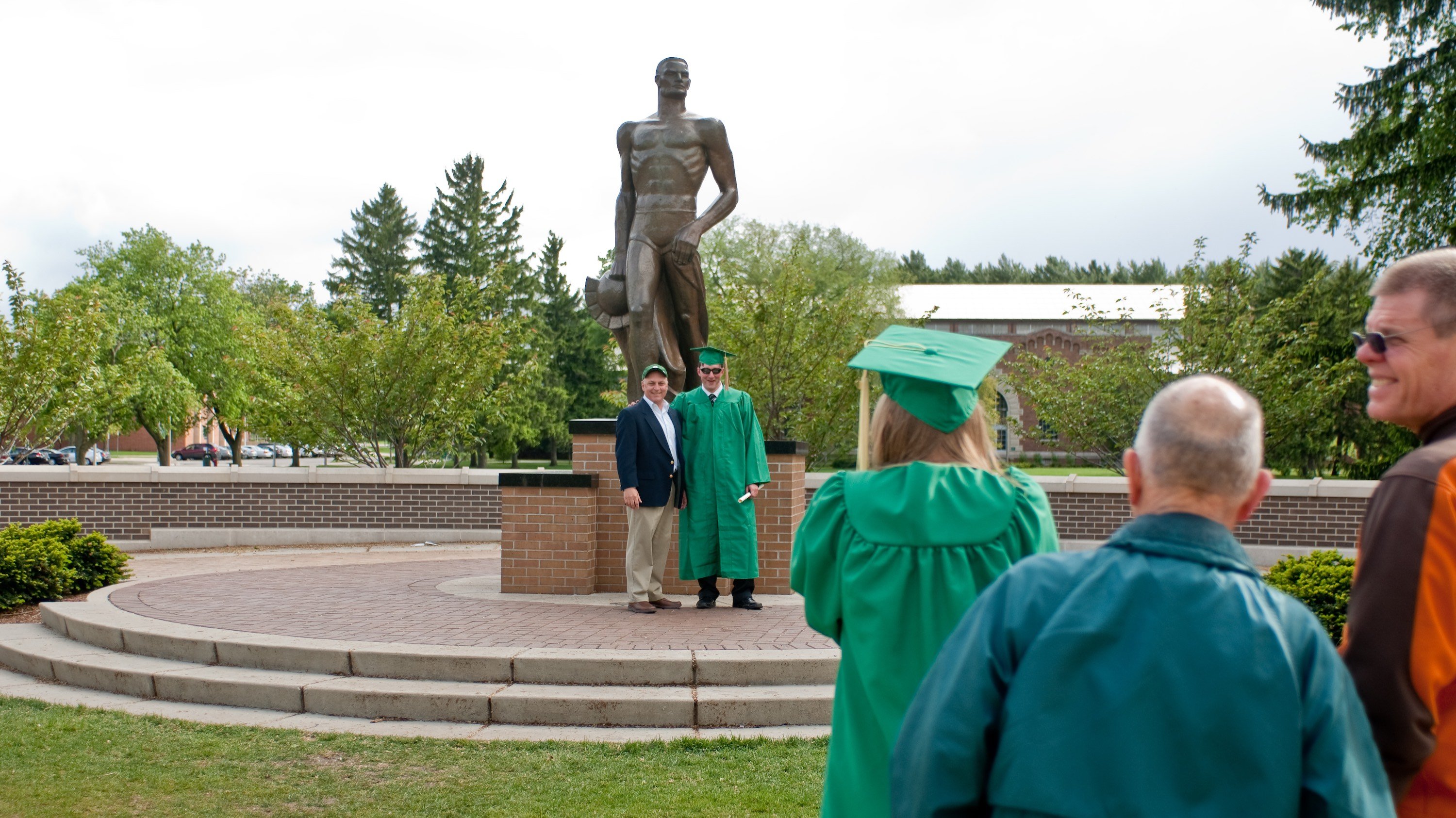 An MSU graduate in cap and gown poses with a family member or friend in front of the Spartan Statue as another graduate snaps a photo. From 2010.