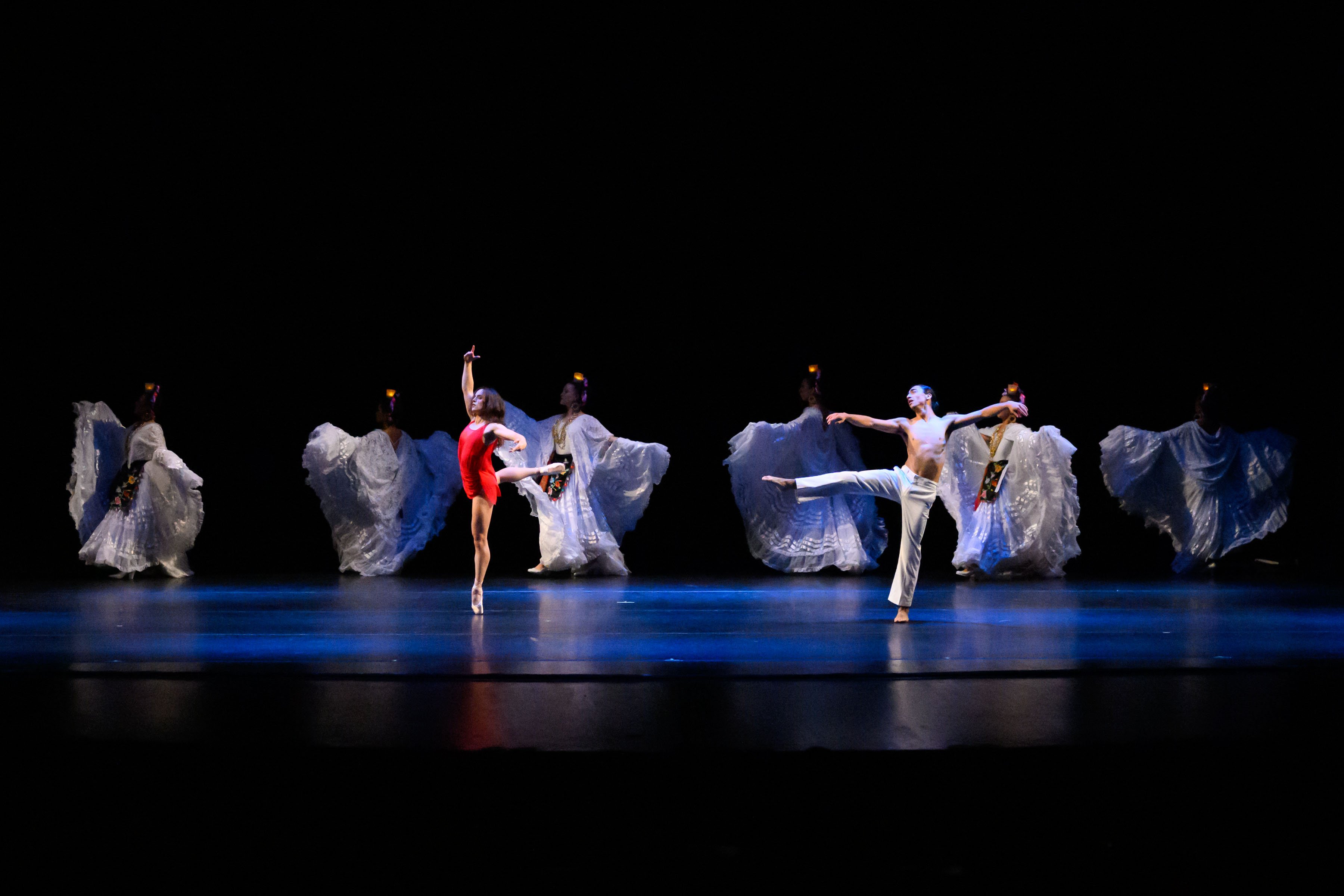 folk costumed dancers in a line across stage with ballet dancers in red and white in the foreground