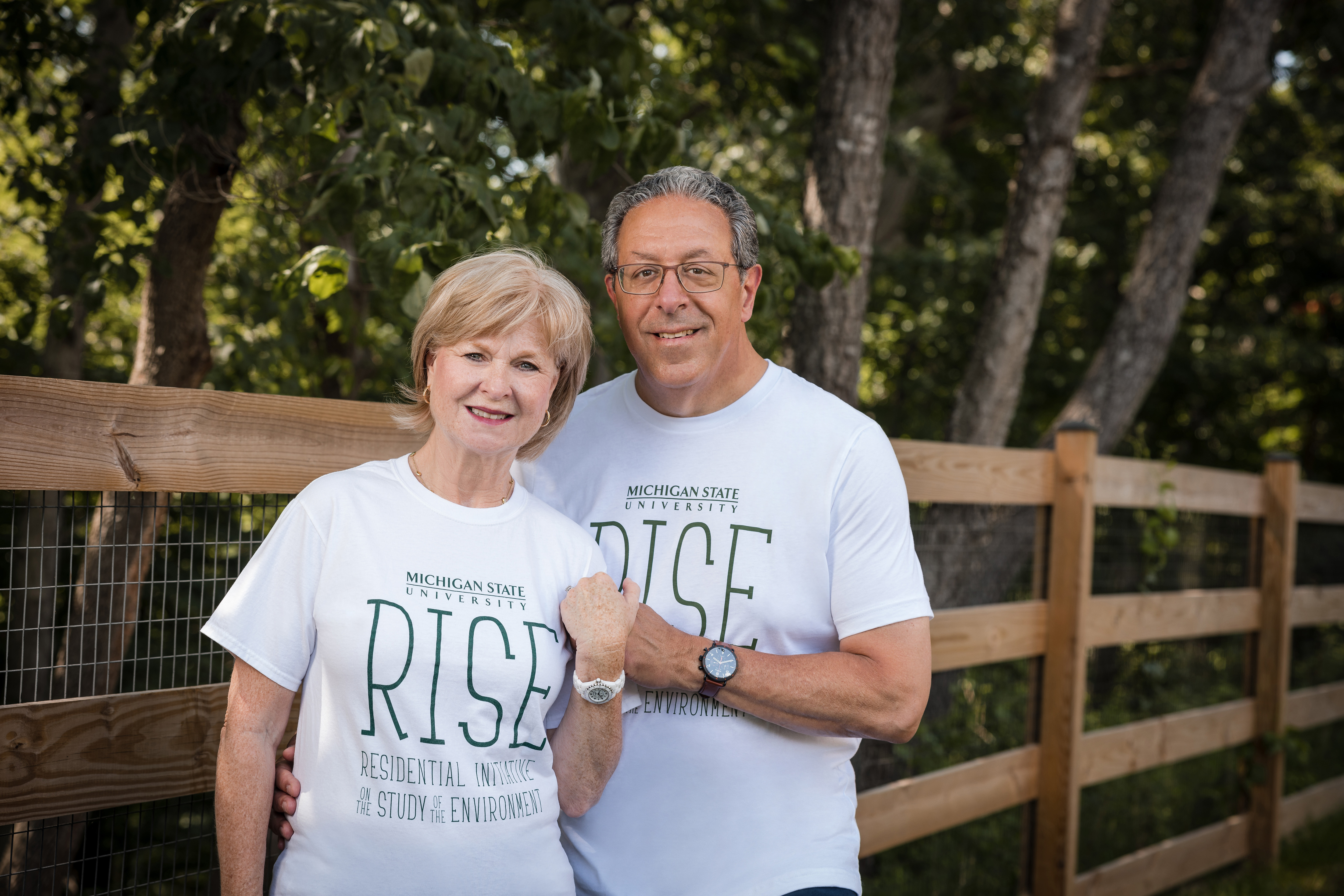 Sue and Gary Farha, both alumni of Michigan State University’s Eli Broad College of Business, are outside in front of a wooden fence in front of trees.