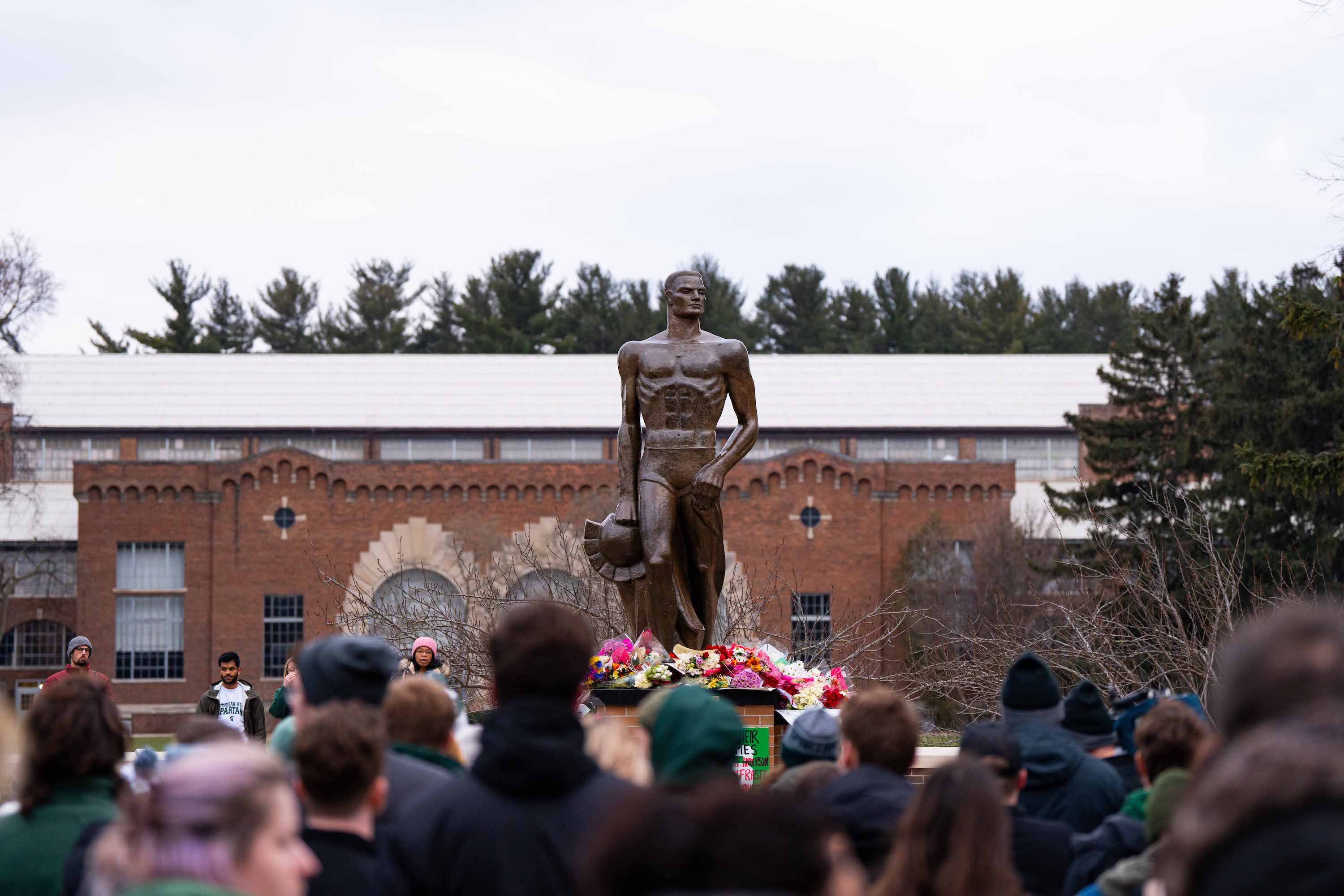 A crowd gathers in front of the Spartan Statue with flowers at the base.