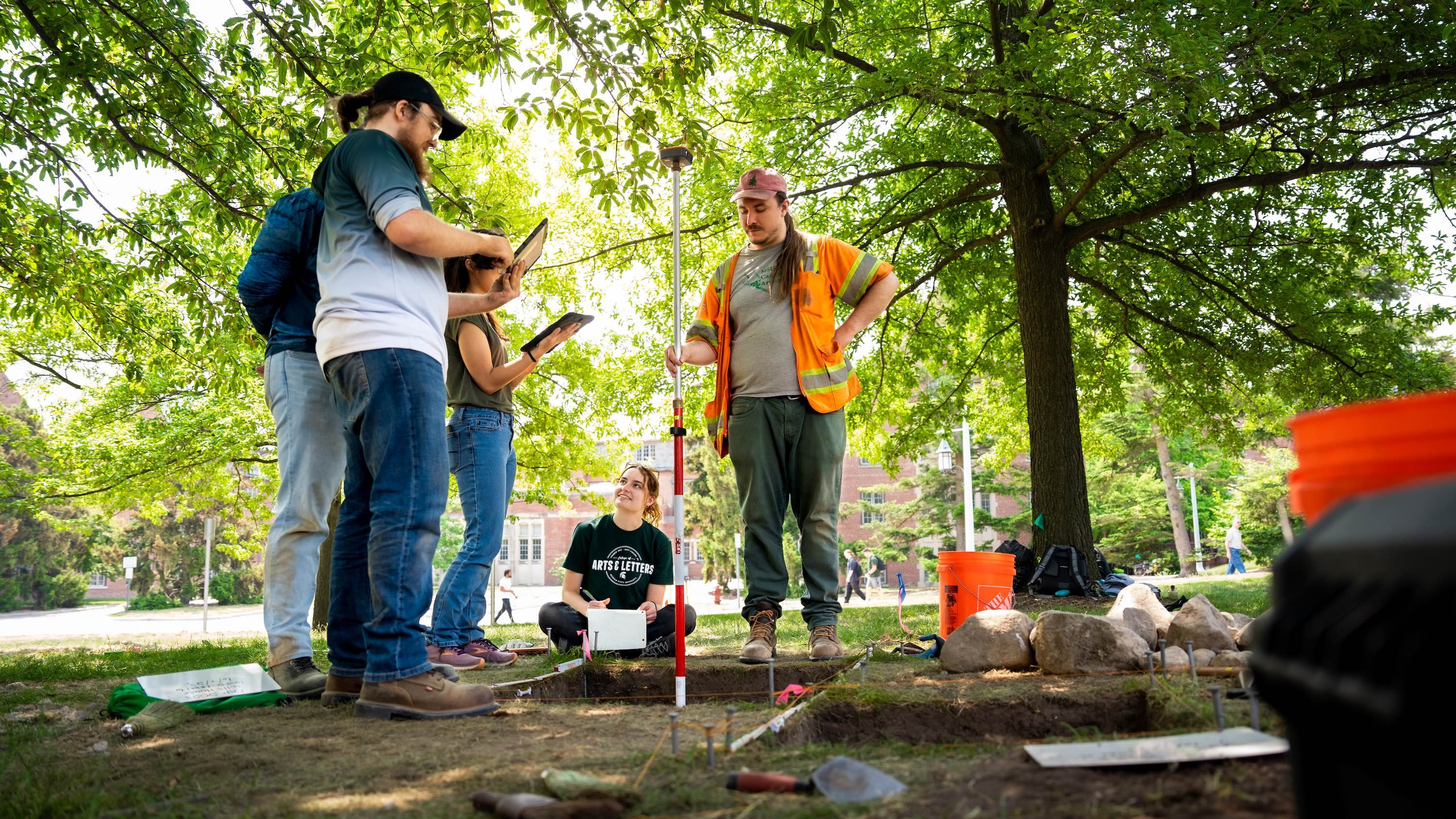 A person holds a long rod in one of two square holes dug into the grass. Three others take notes on paper or iPads.