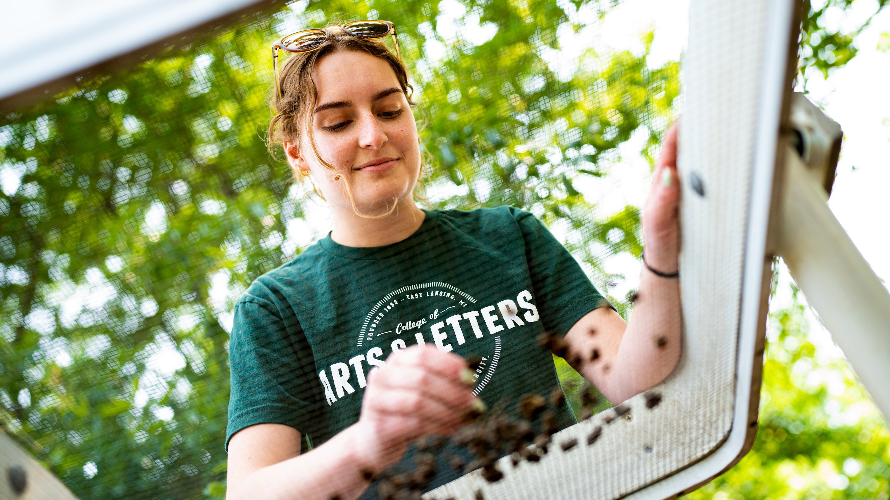 A young woman uses a large mechanism to sift through soil. The image is taken from underneath the large sifter.
