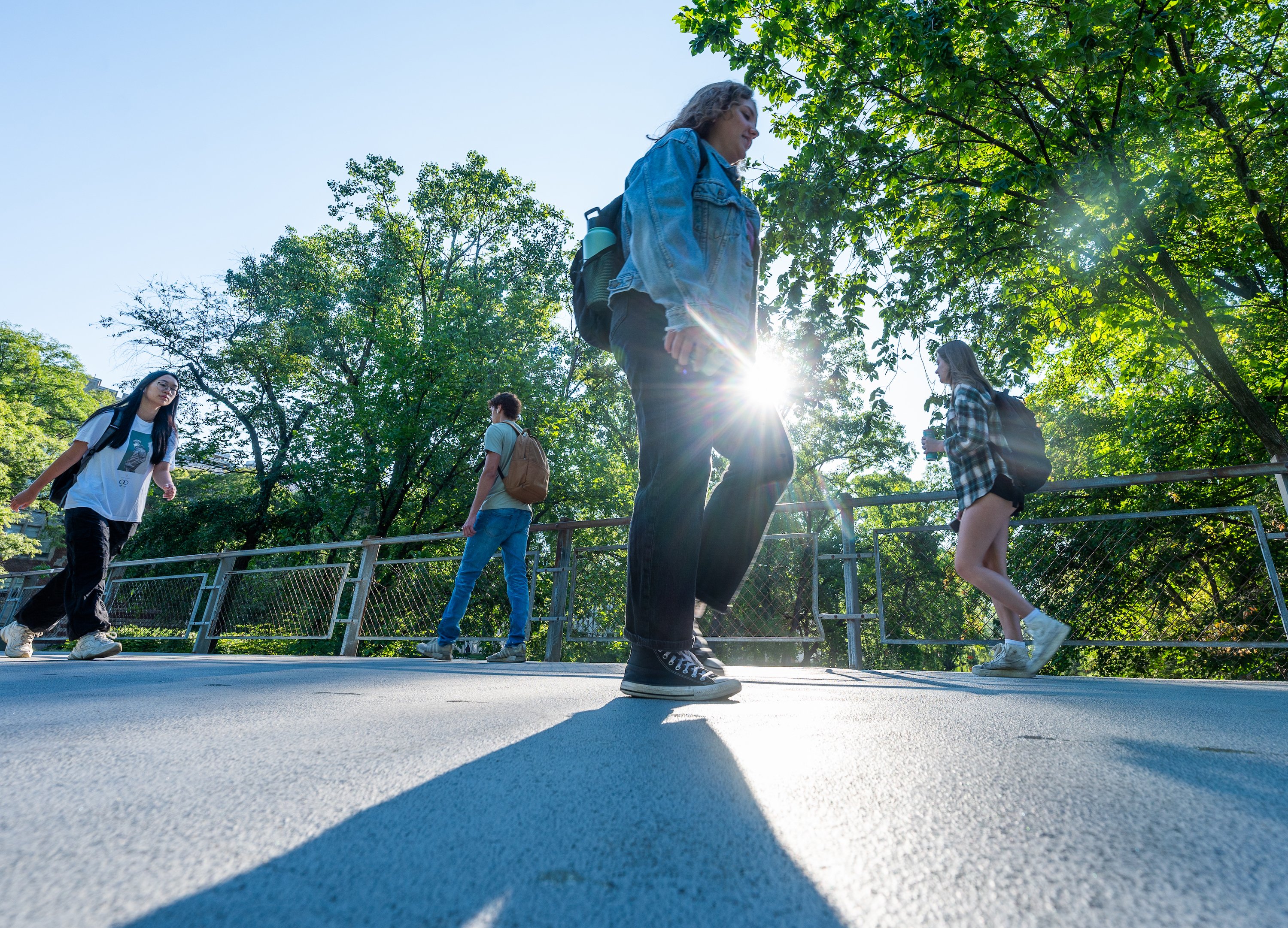 Students walking outside on a foot bridge. Lush, green-leaved trees are behind as they walk.