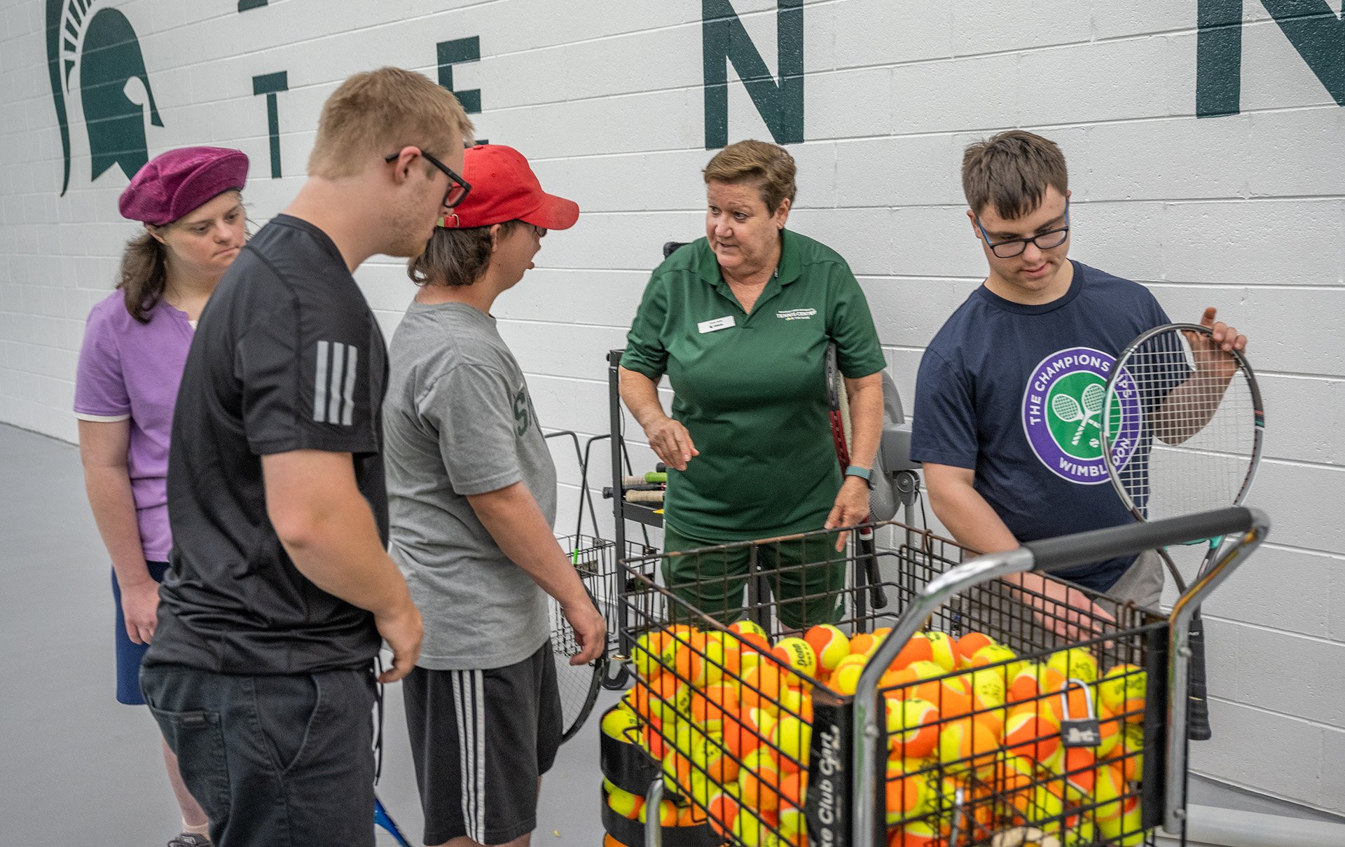 Participants gather around the tennis coach and a cart of yellow and orange tennis balls