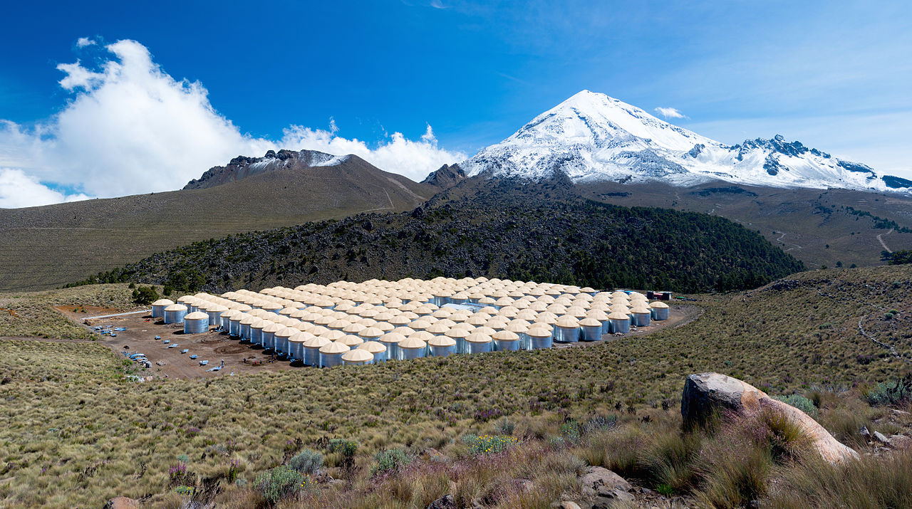 A photograph of the High-Altitude Water Cherenkov Observatory in Mexico. Hundreds of tan-domed steel water tanks sit beneath a snow-capped peak in the background.
