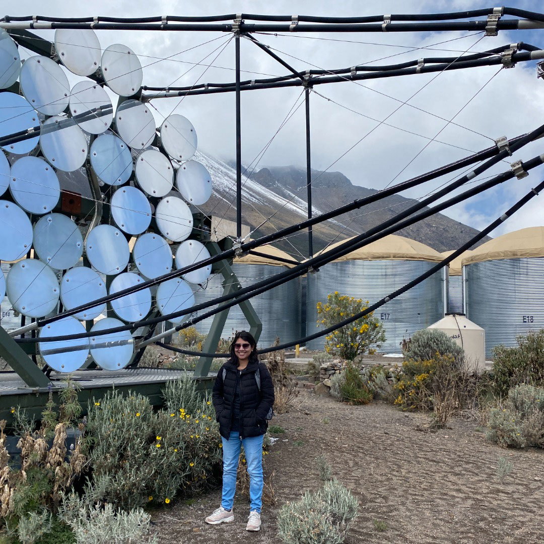 Mehr Un Nisa stands in front of some of the water tanks at the High-Altitude Water Cherenkov Observatory. A snow-capped mountain peak can be seen in the background, disappearing into the clouds.