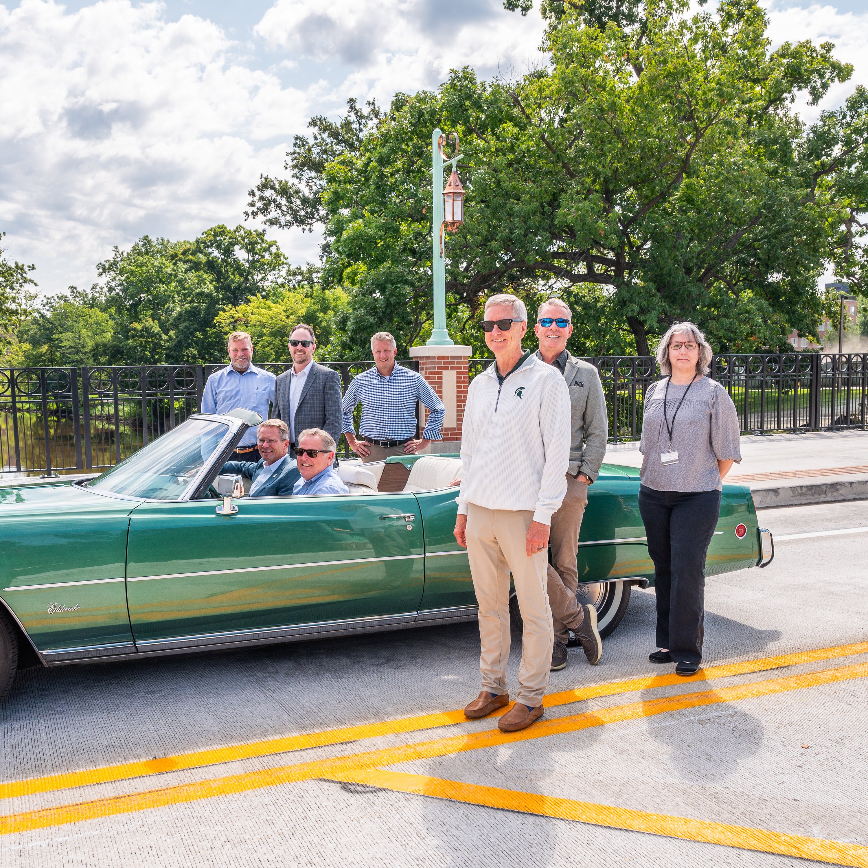 A group of individuals pose around a green car on a roadway.