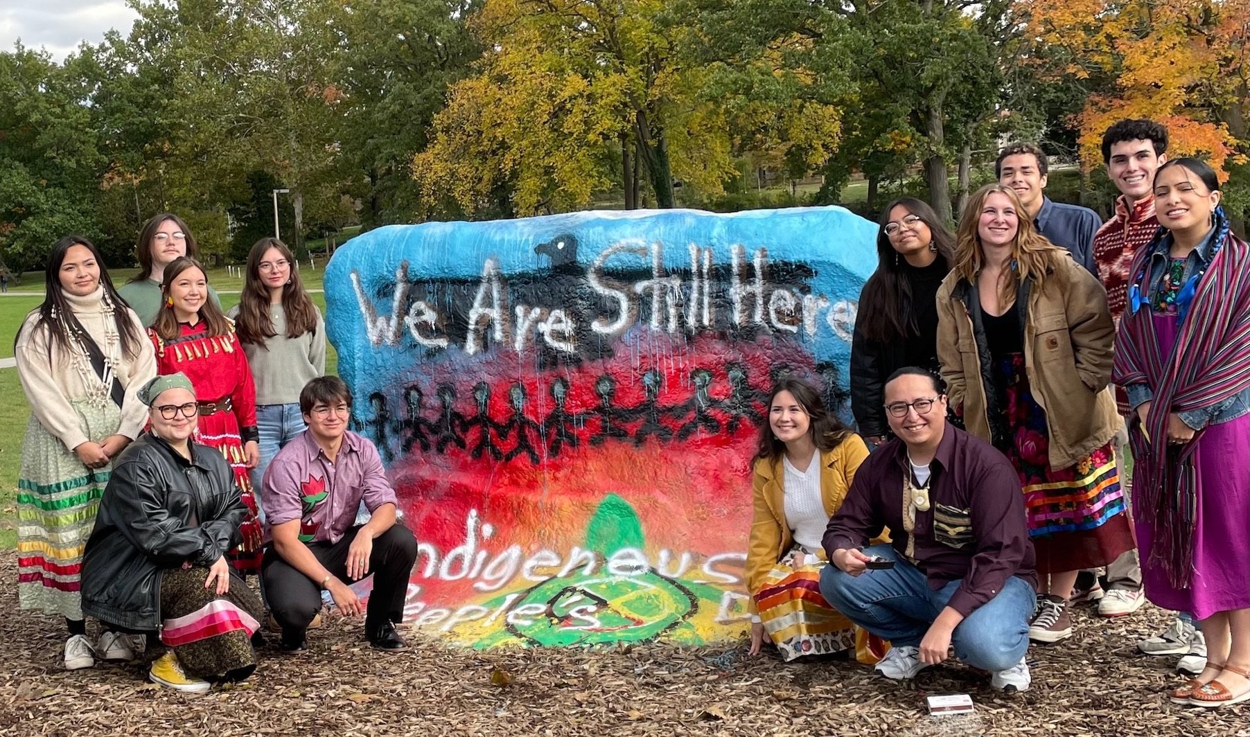 Individuals stand near the Rock, where text says: We are still here. Indigenous Peoples' Day 2024