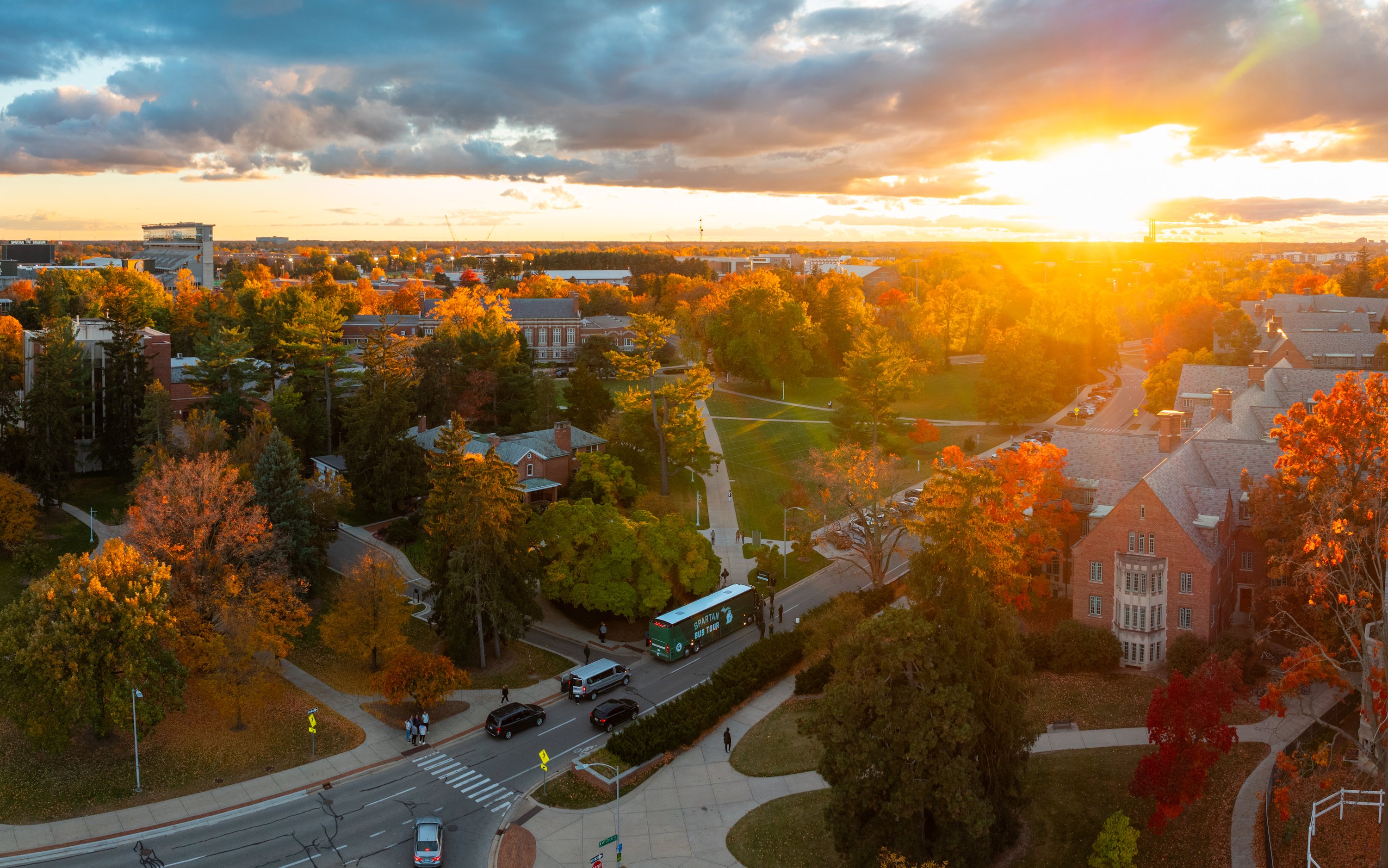 A drone image of the Spartan Bus Tour bus driving in front of Cowles House on MSU's campus set against a fall sunset.