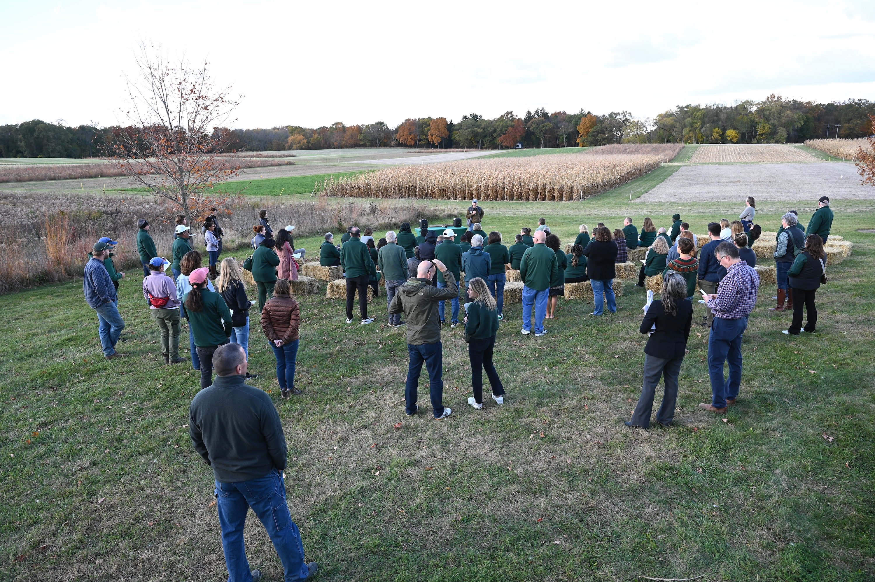 Individuals stand outside on grass with fields in the background.