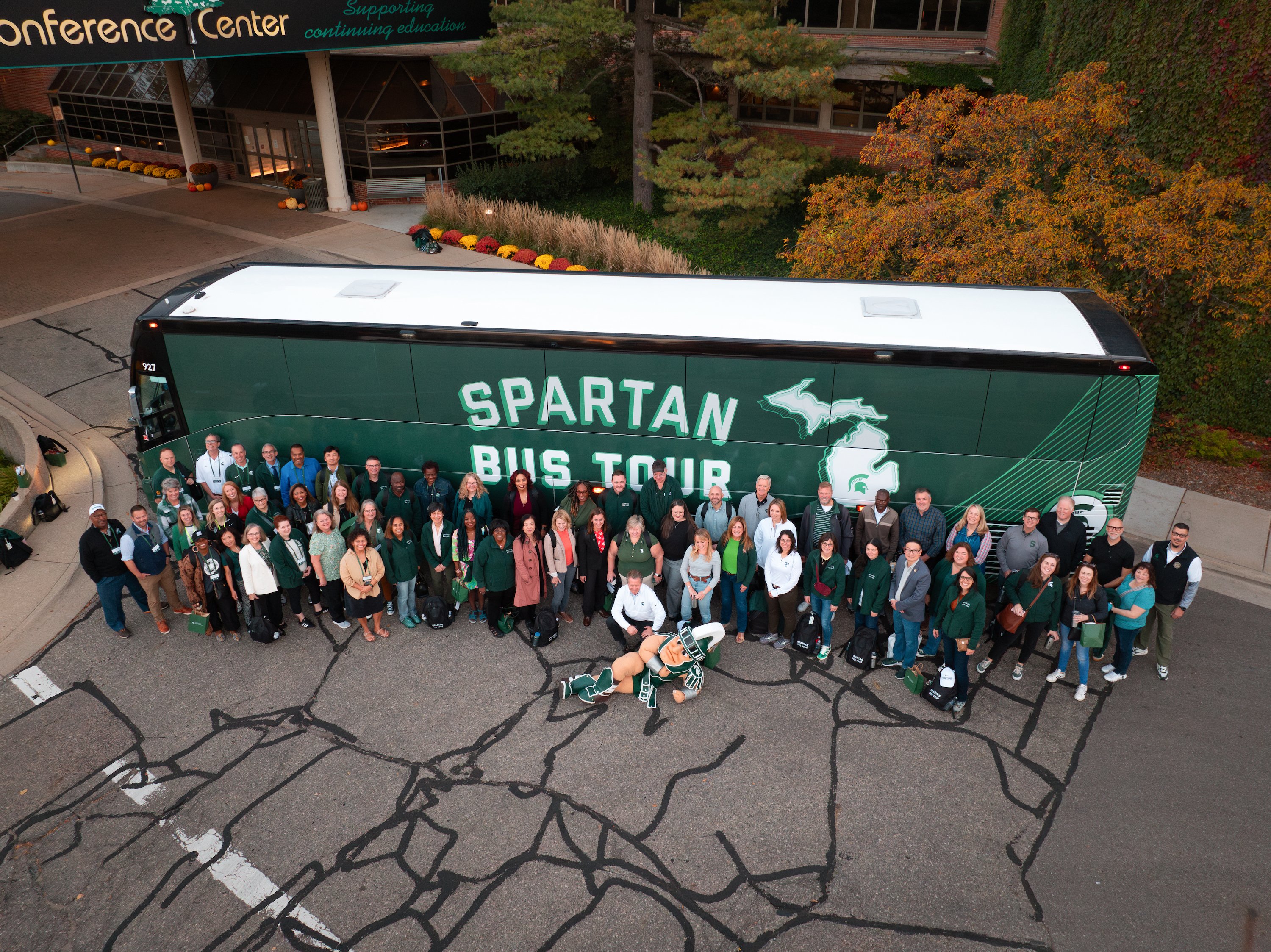 A group of people, and Sparty the mascot, pose in front of the Spartan Bus Tour bus at the Kellogg Hotel and Conference Center.