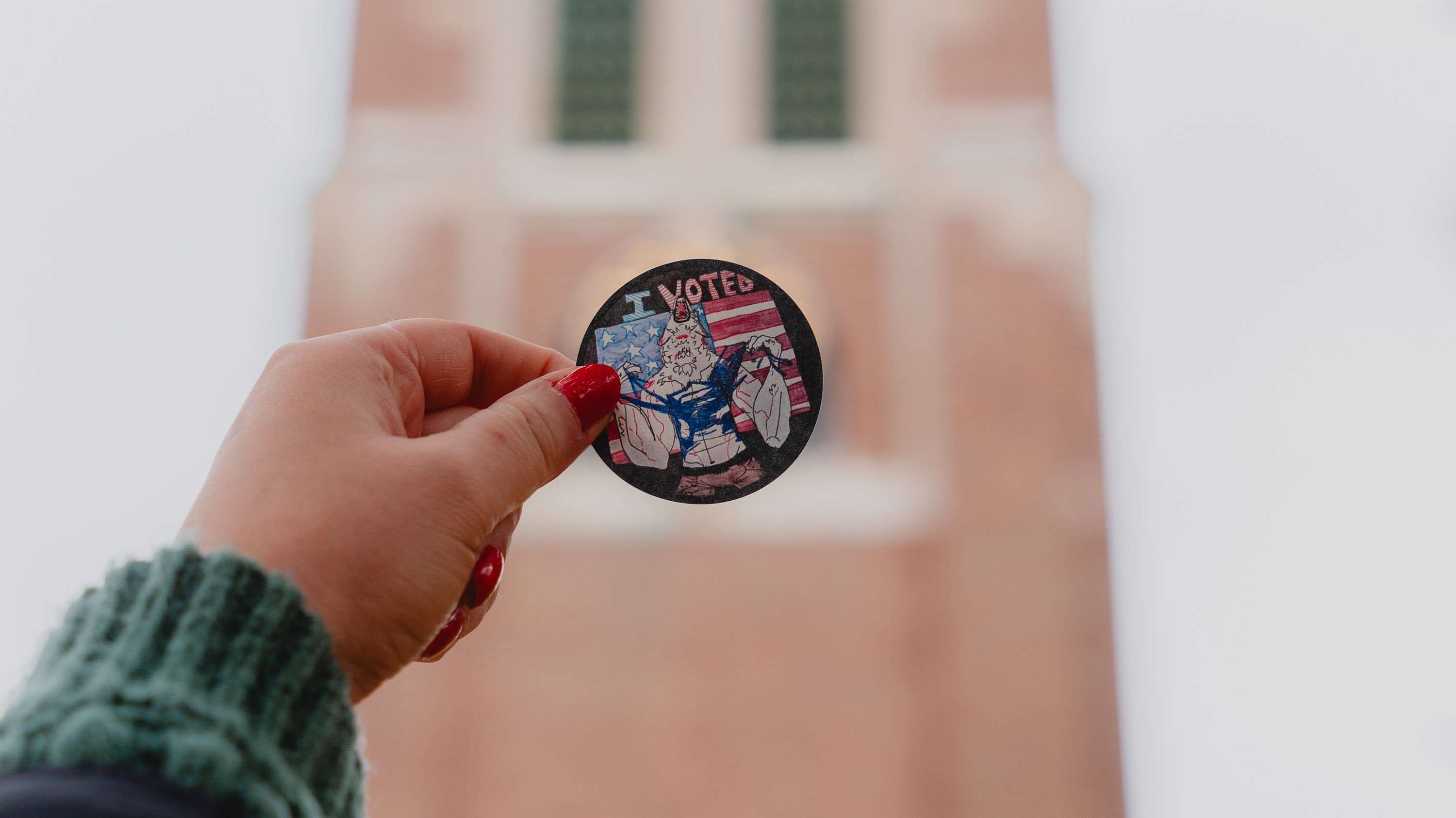 Hand holding a werewolf 'I voted' sticker in front of Beaumont Tower
