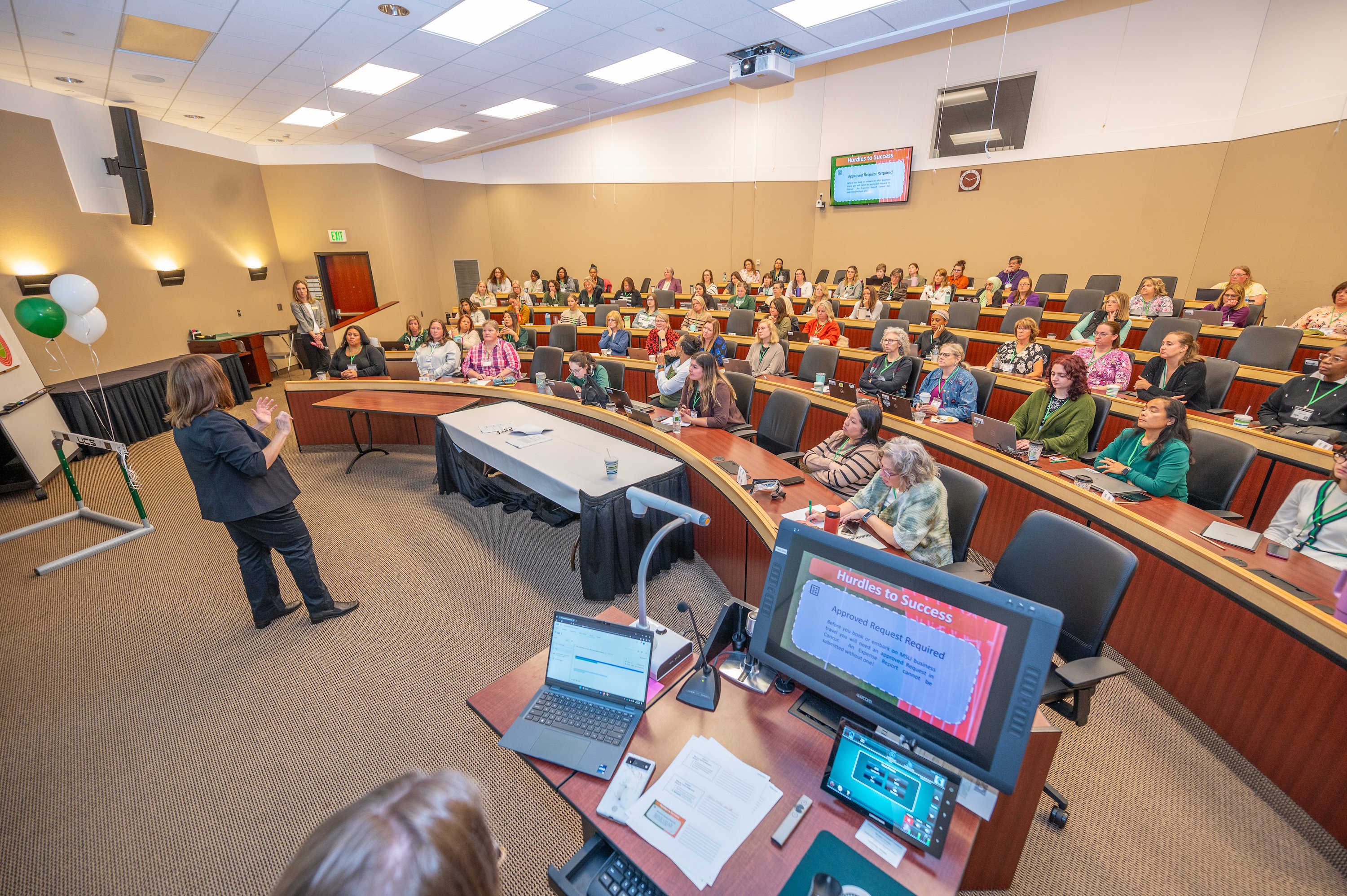 A presenter stands at the front of a room filled with participants sitting in seats.