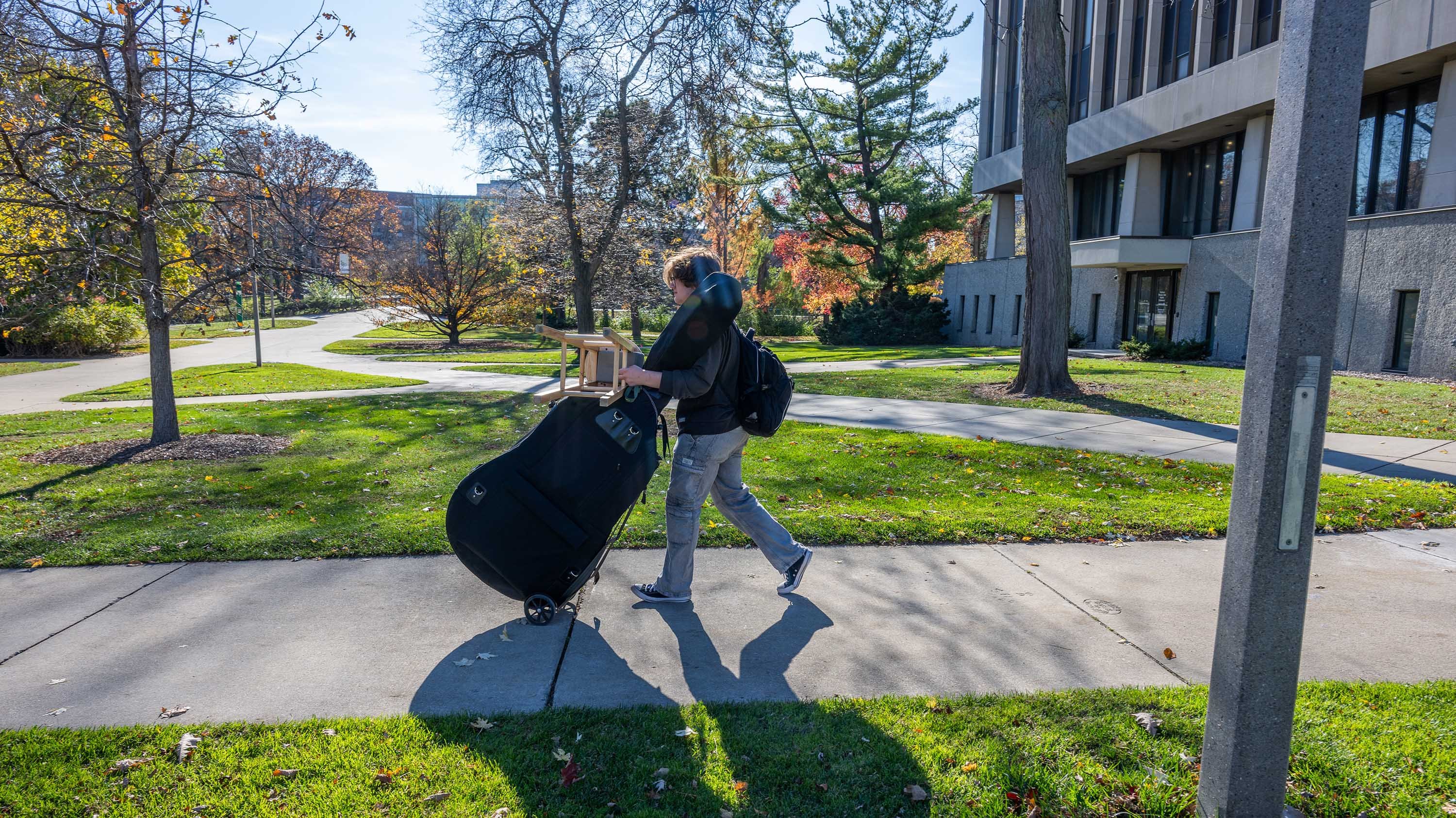 Student carrying double bass and stool across campus