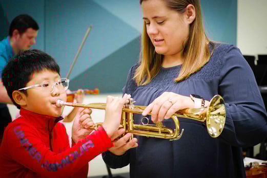 An adult helps a child hold the trumpet while the child plays the trumpet.