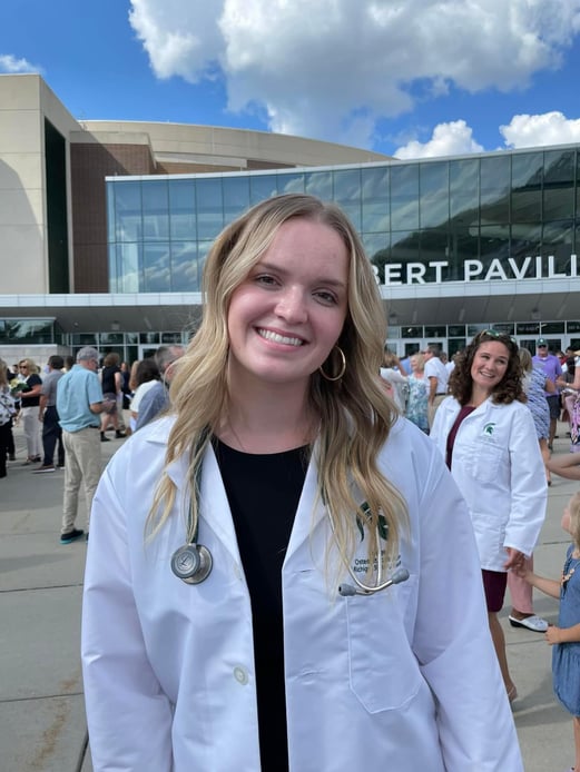 A medical student in a white coat standing outside of the Gilbert Pavilion of the Breslin Center.