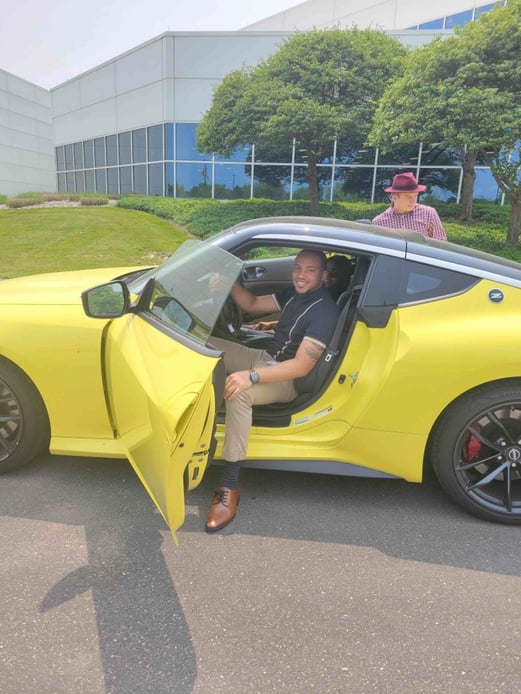 A person sits in a yellow car with the driver's side door open, while an another individual is in the background.