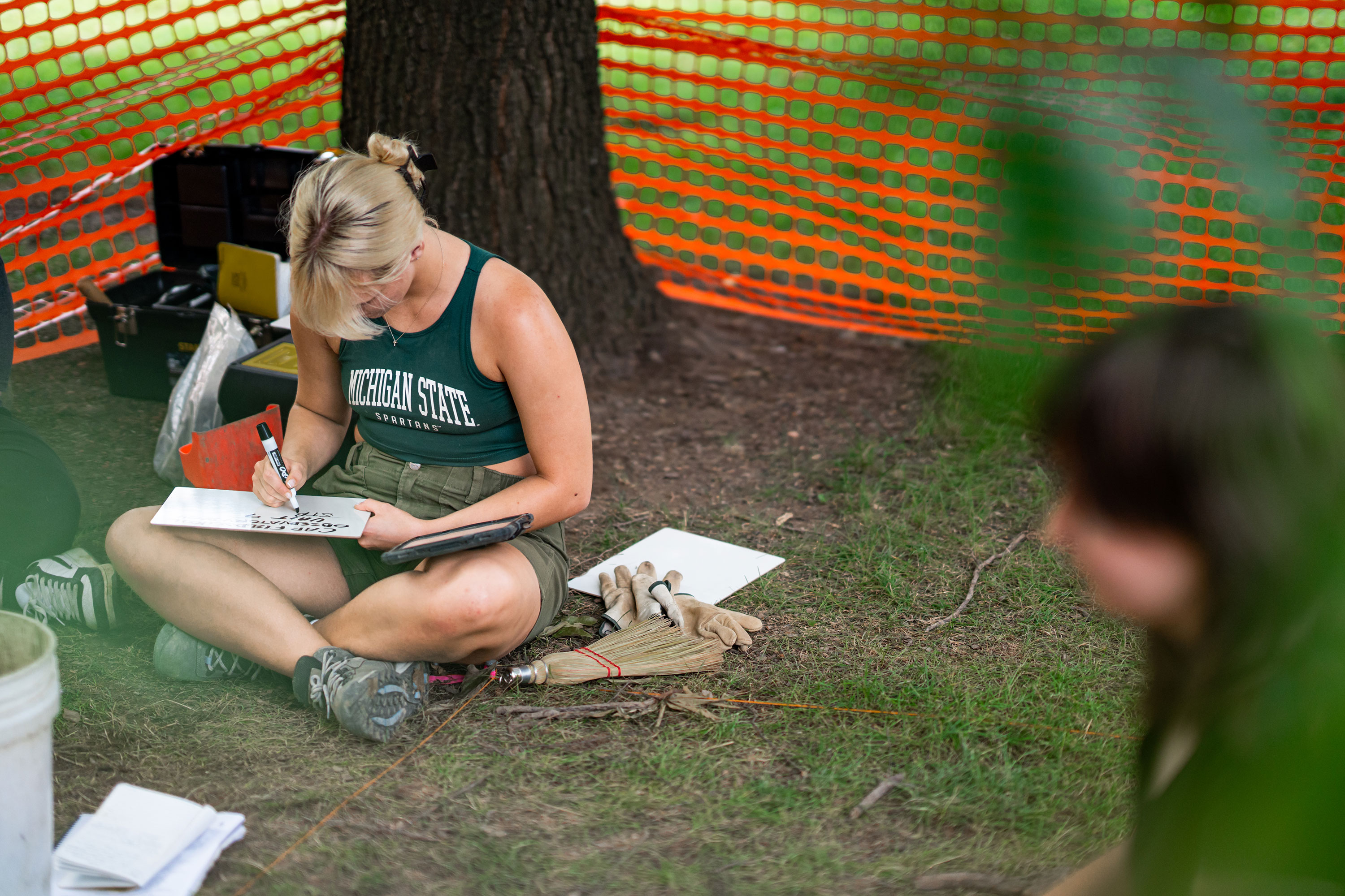 A woman sitting in the grass and writing on a dry erase board.