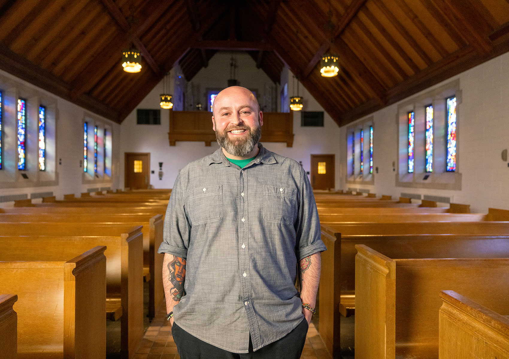 A man standing in the center aisle of a chapel. Sun is shining through the stained glass windows.