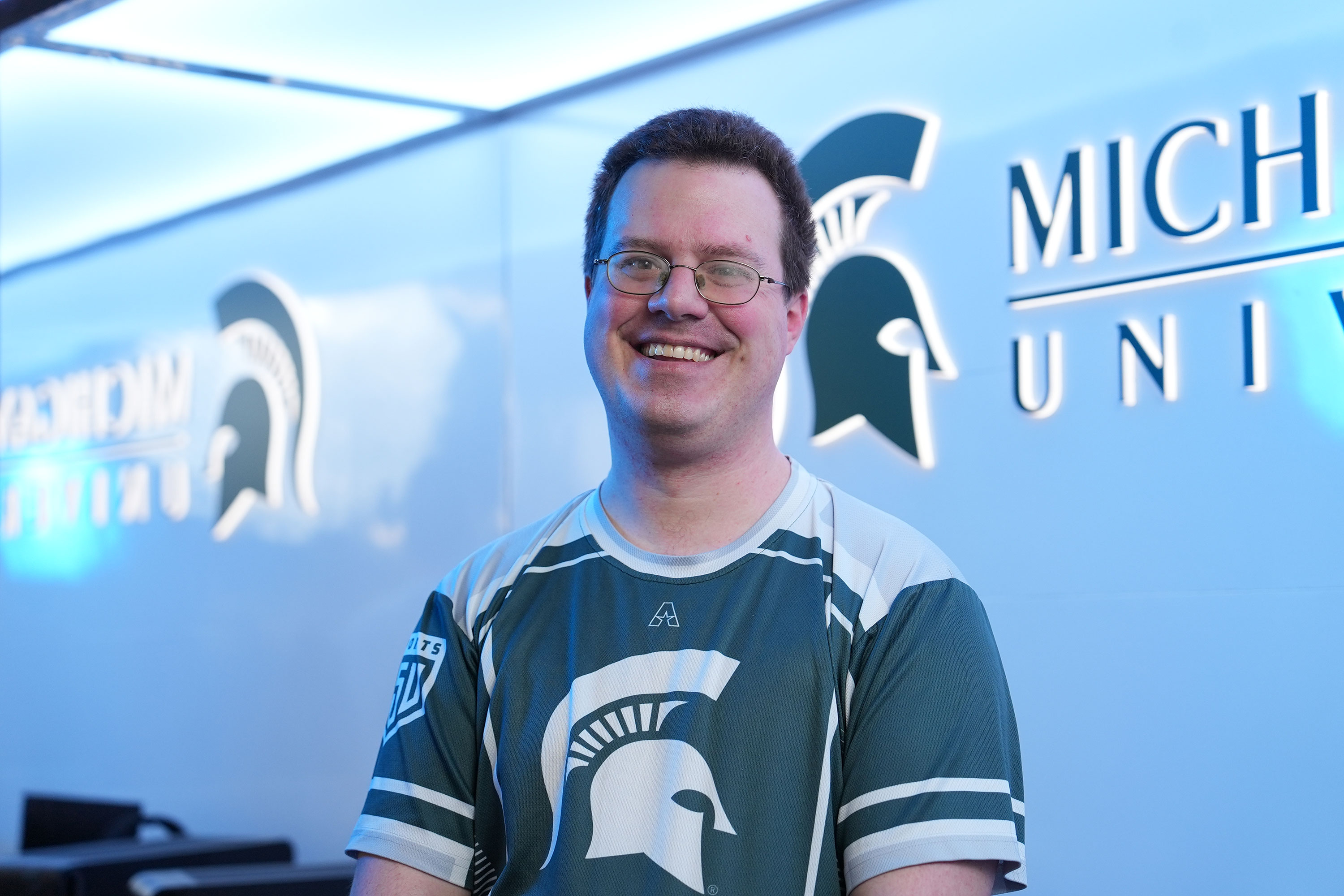 A person posing for a portrait in a silver room. A green Michigan State University Spartan head logo is on the wall behind them.