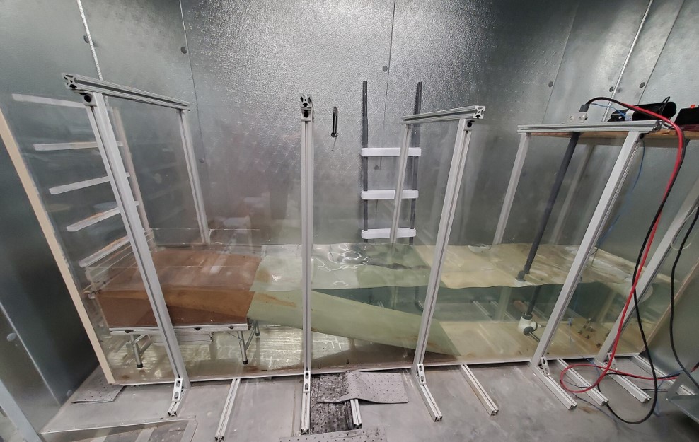 Side profile of the wave tank device. To the left of the photo is the beach ramp and the sediment beach. On the right side of the photo is the plunging wave generator and a heating element. When the tank is in use, cameras are mounted to the overhead aluminum struts. The wave tank is located within a large walk-in freezer.