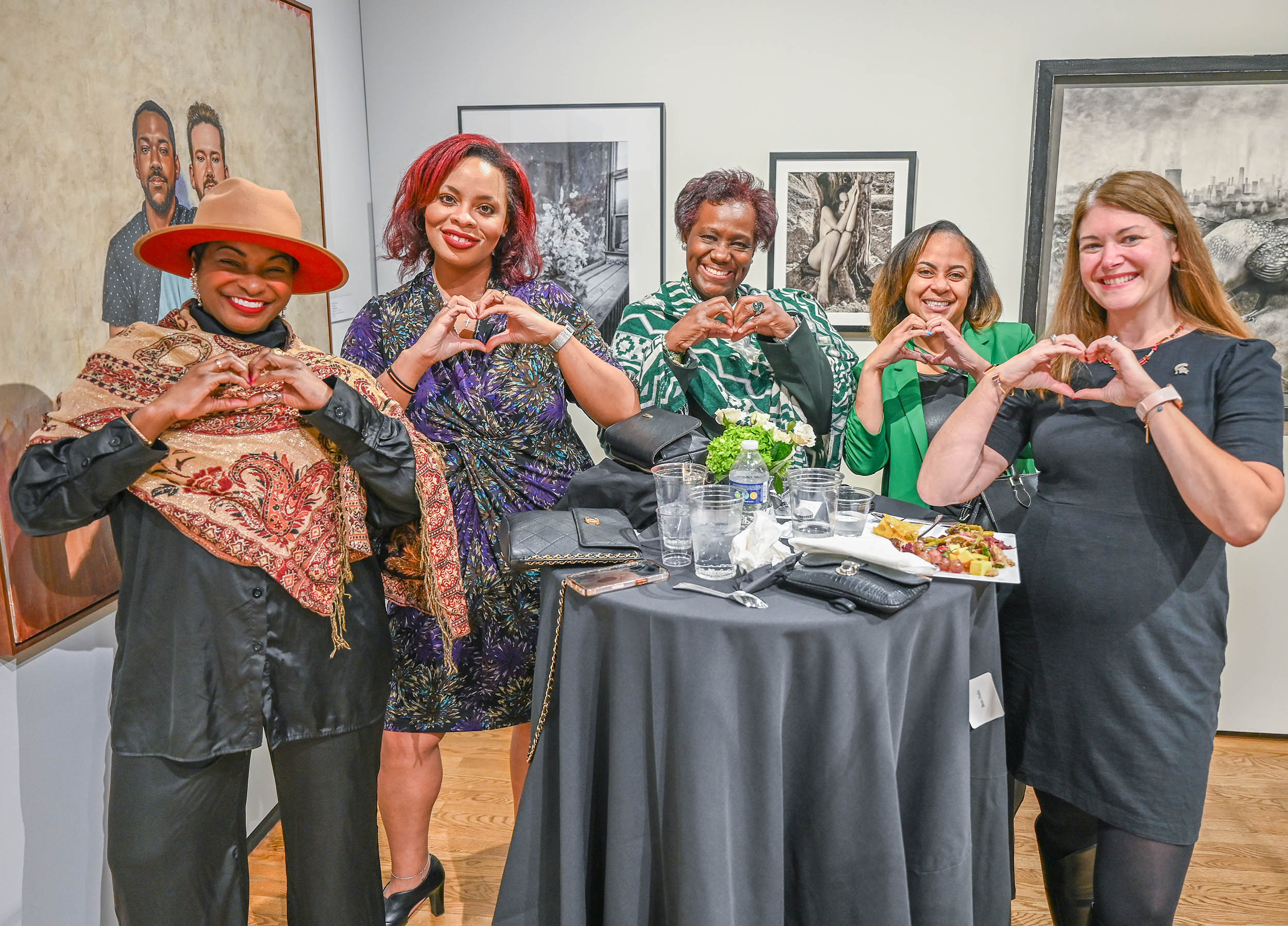 A group of women pose for a picture in an art gallery. They are standing around a table and making a heart shape with their hands.