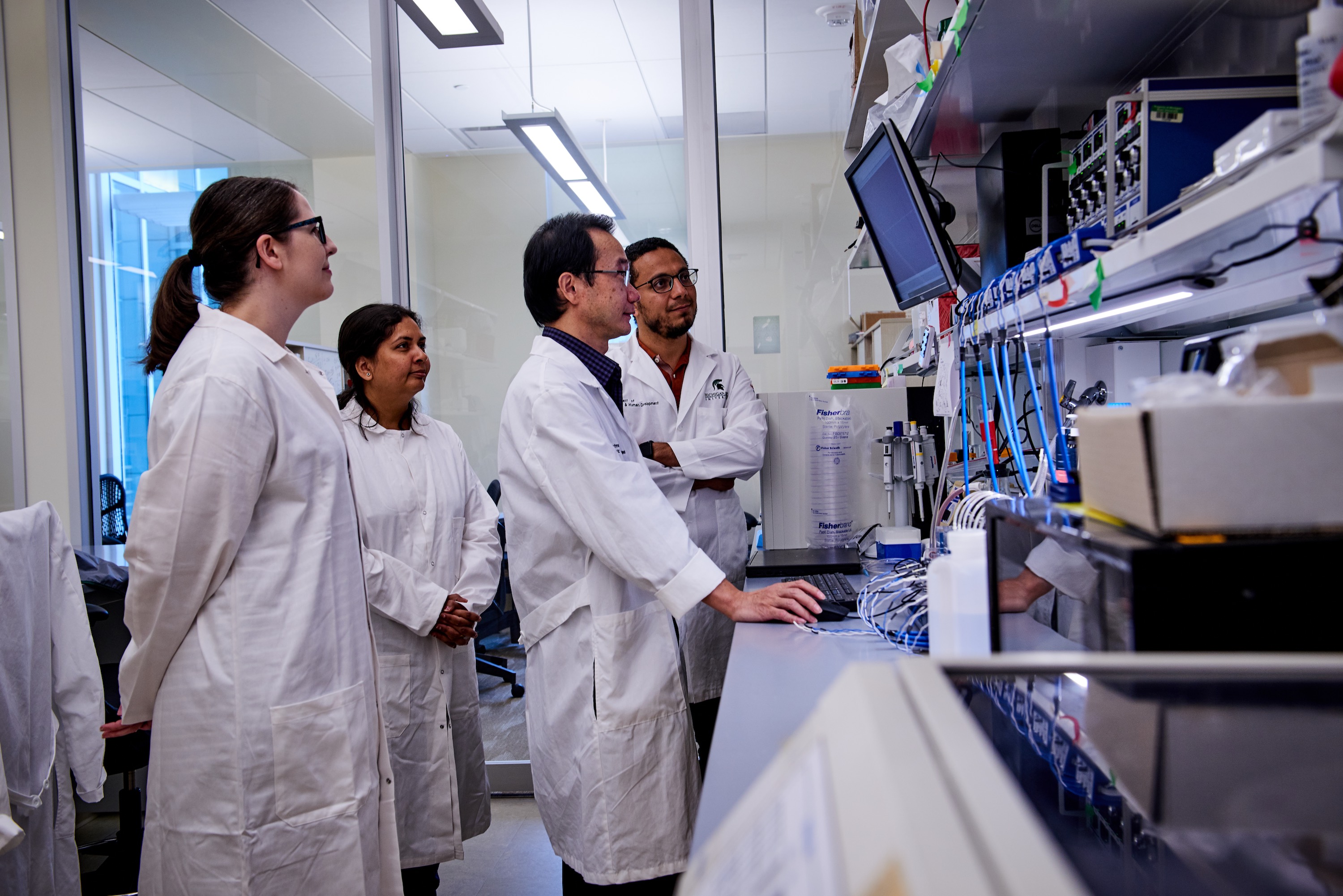 A group of four scientists in white lab coats look at a computer screen in a lab