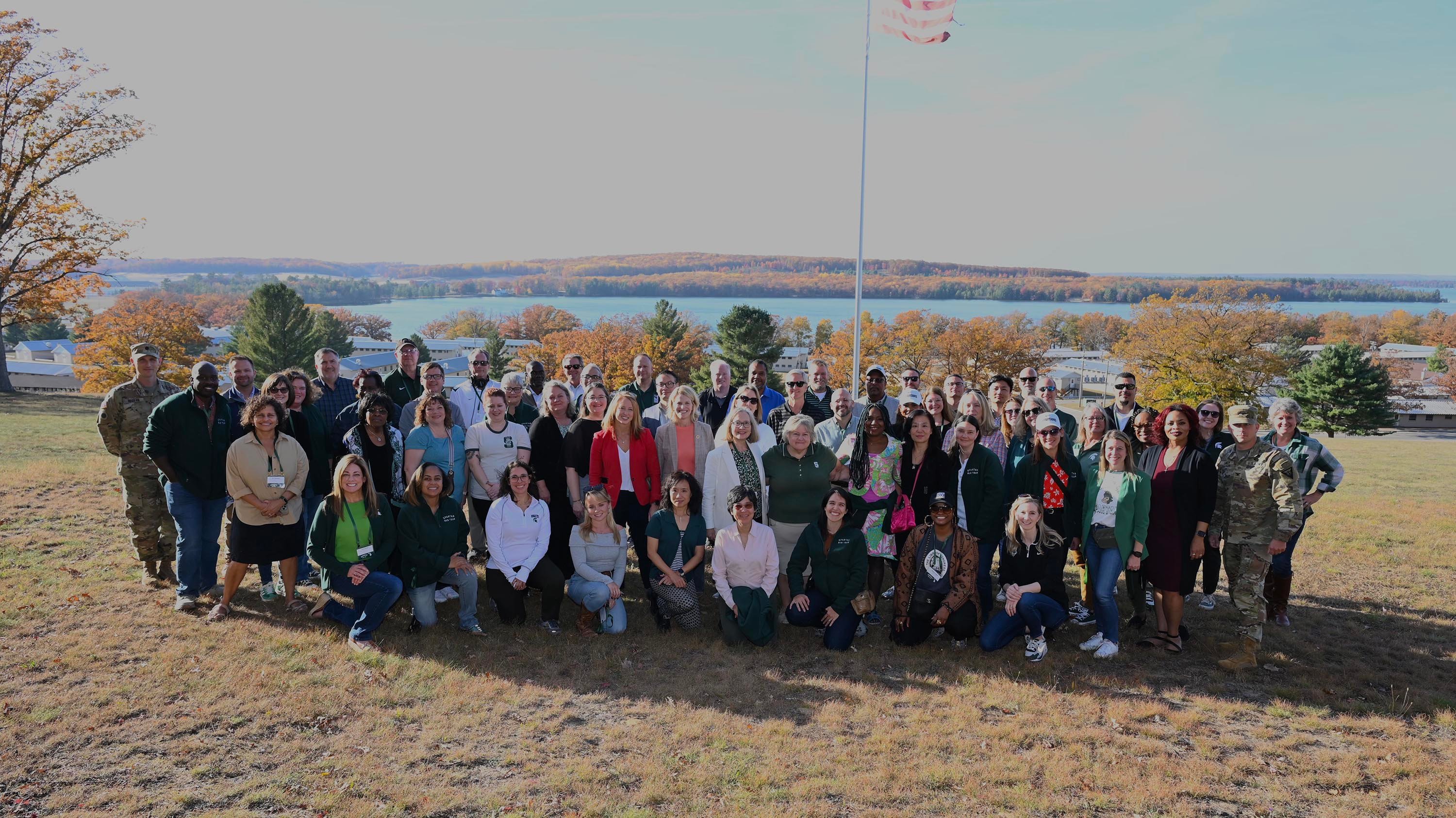 A group of people posing for a photo in the fall. Behind them, you can see a flagpole with the American flag, lakes and trees in various shades of orange, yellow and red.