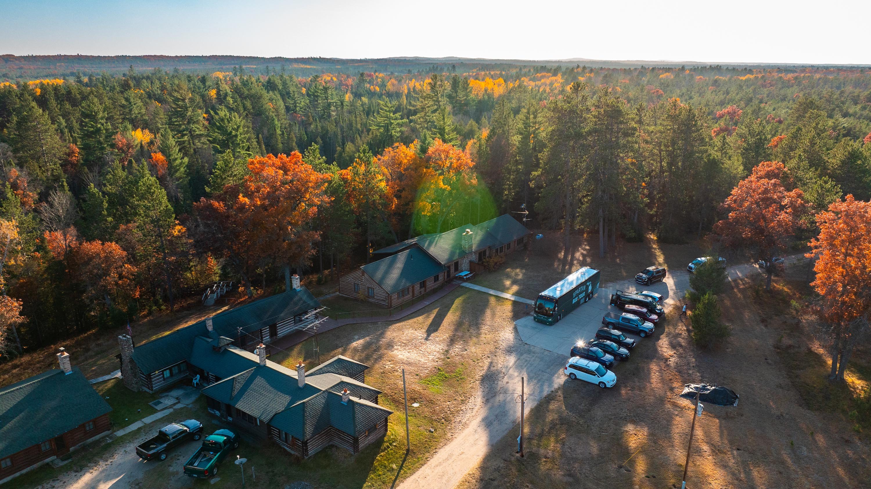 An aerial photo of a cabin in the fall. There are multiple vehicles in front of the cabin, including a large green bus.