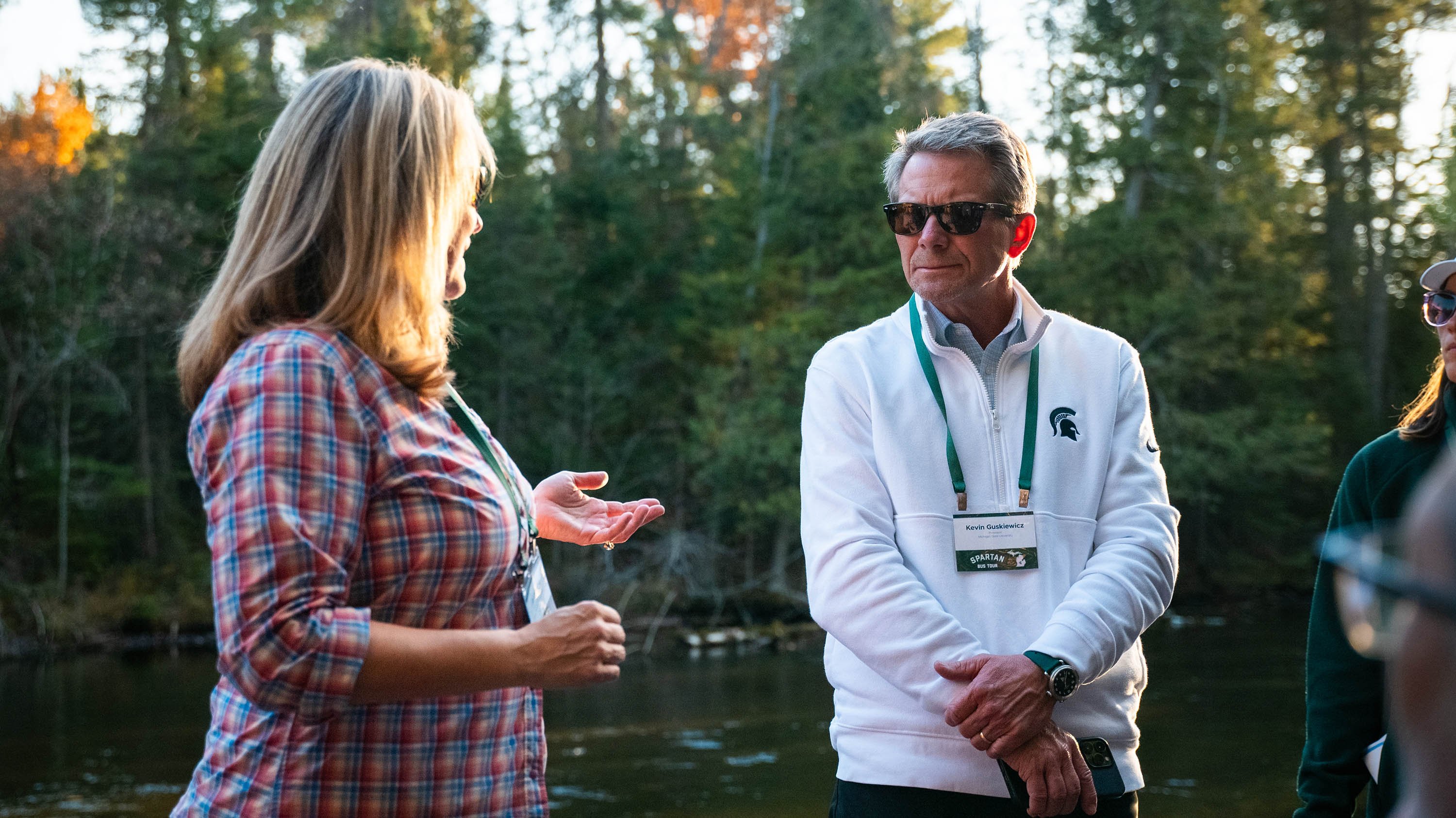 Two people talking near a river. There are pine trees in the background.