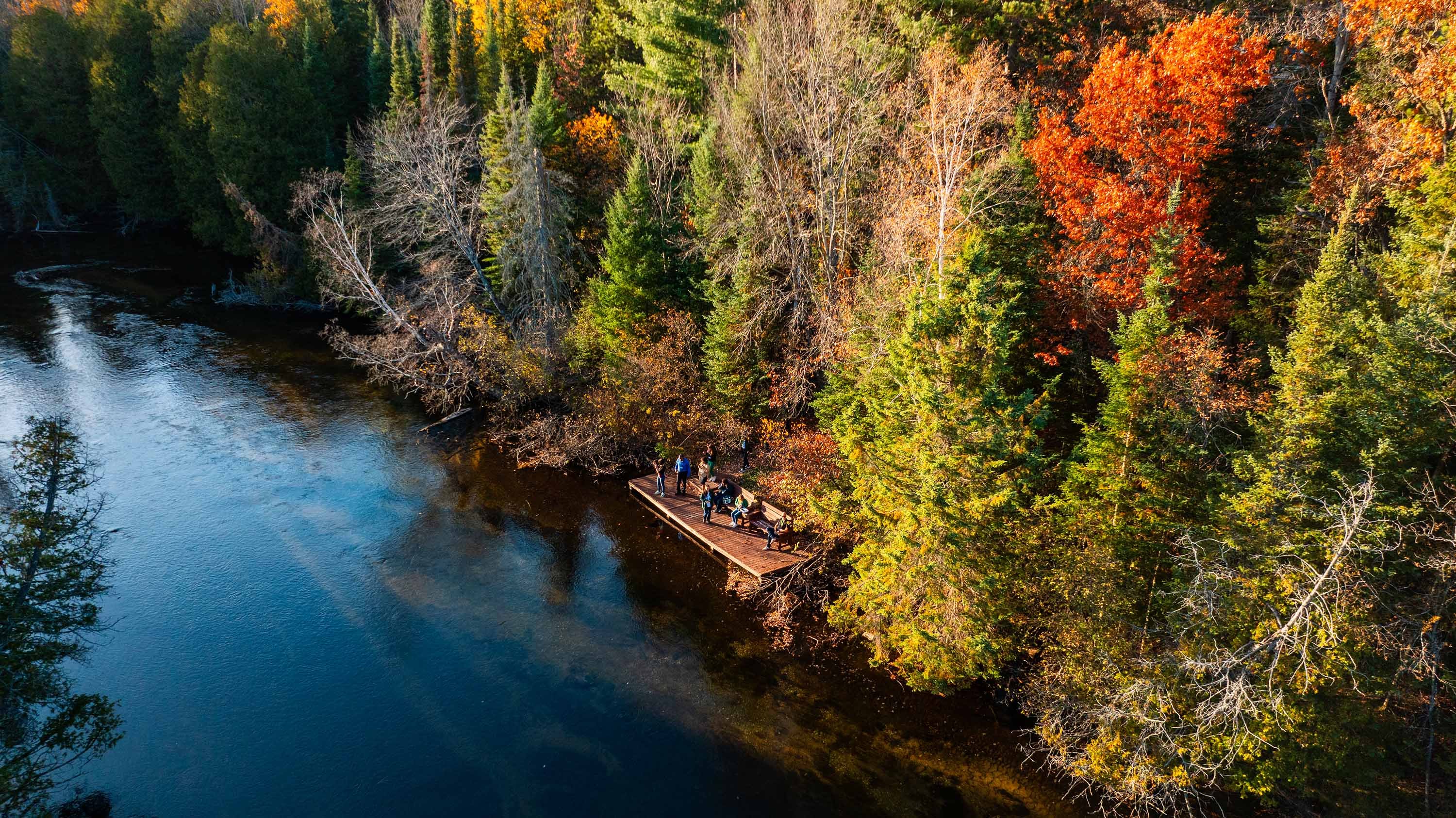 An aerial view of a river in the fall. There is a dock on the shore of the river where there are people standing.